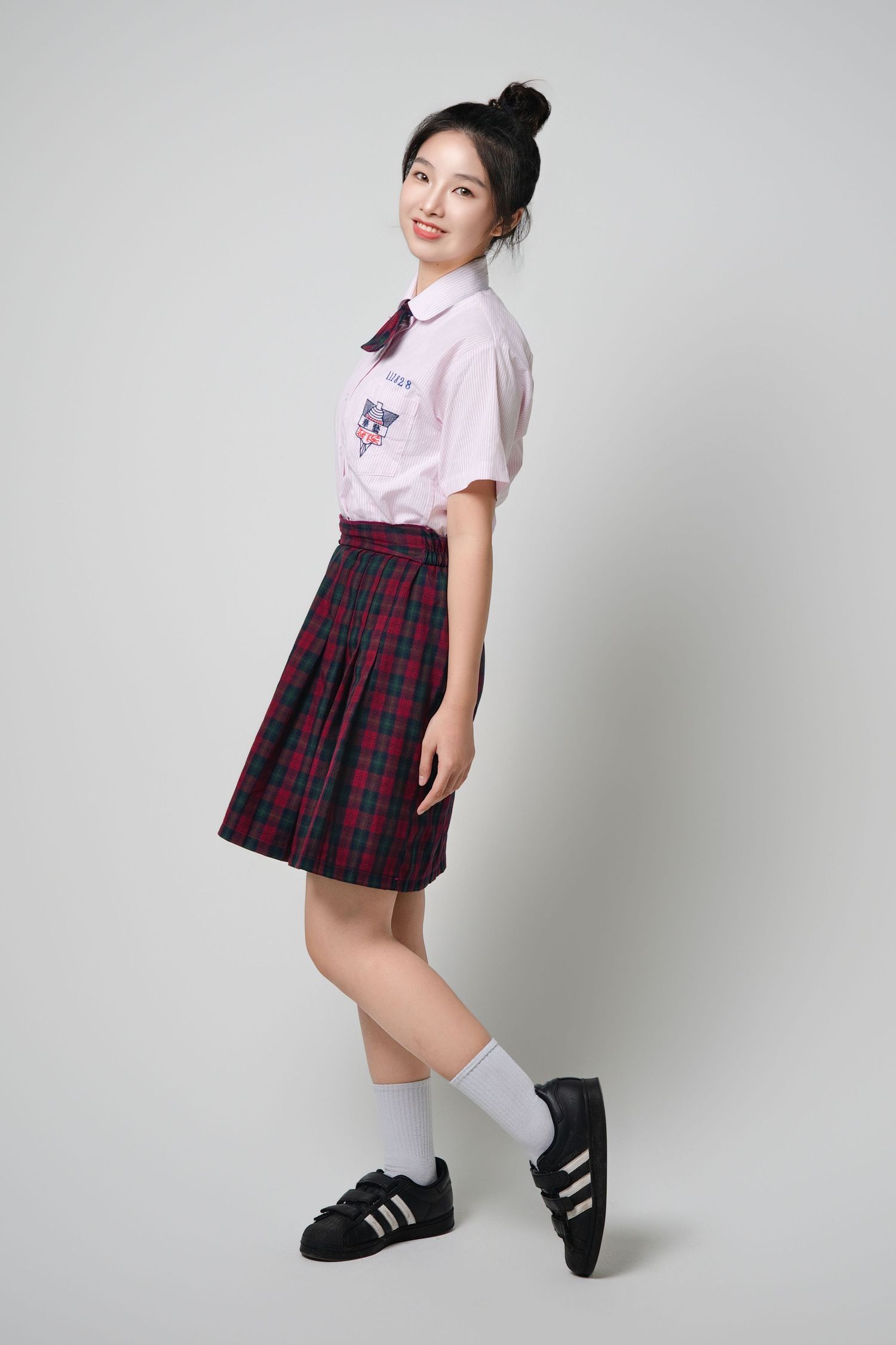 Student wearing school uniform with pink shirt, plaid skirt, white socks and black sneakers poses against grey background.