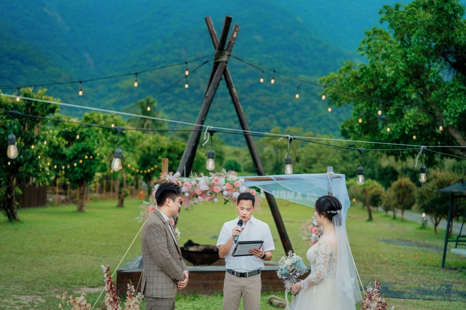 A couple shares a romantic moment under a white tent in a lush green mountainous vineyard setting with string lights.