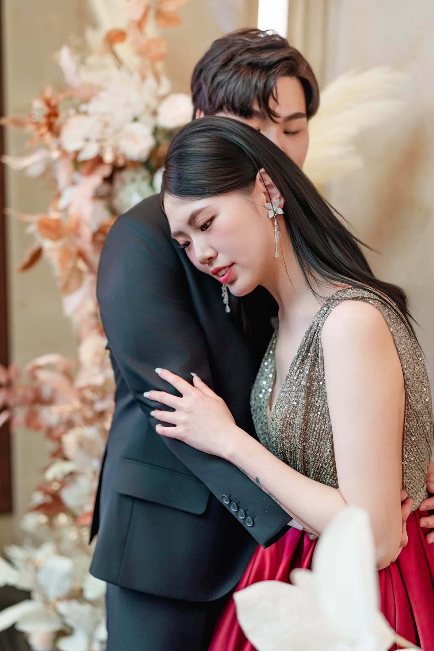A couple in formal attire embrace intimately against a backdrop of soft pink floral decorations.