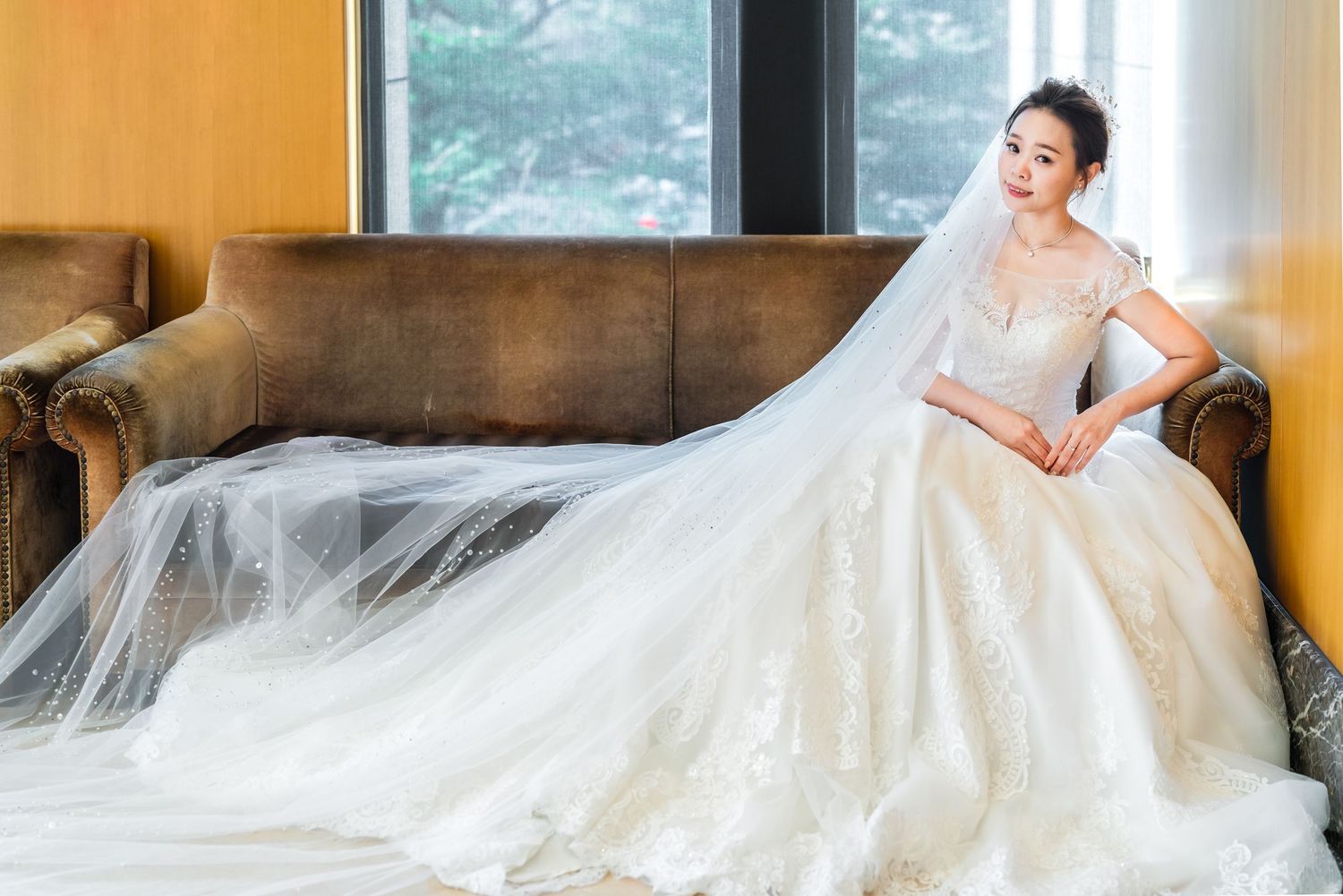 A bride wearing an elegant ivory ball gown with lace detailing lounges on a brown leather couch with a flowing cathedral veil.