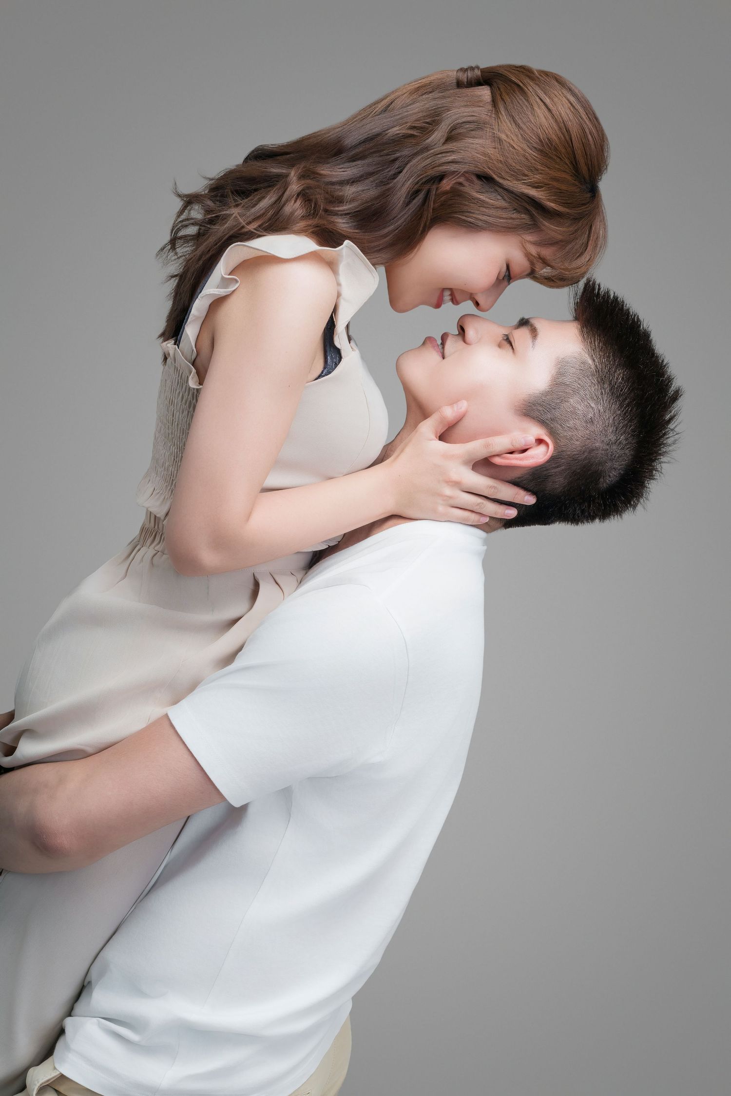 A romantic couple shares an intimate moment in beige and white attire against a gray studio backdrop.
