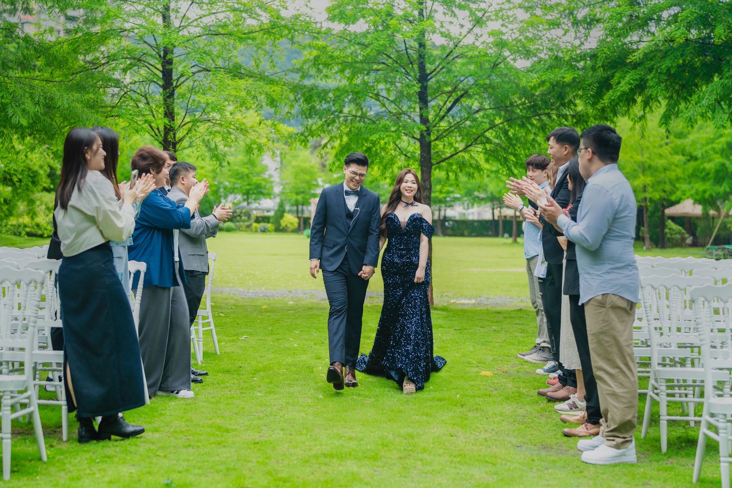 Wedding guests with cameras documenting a couple walking down an outdoor aisle lined with white chairs.