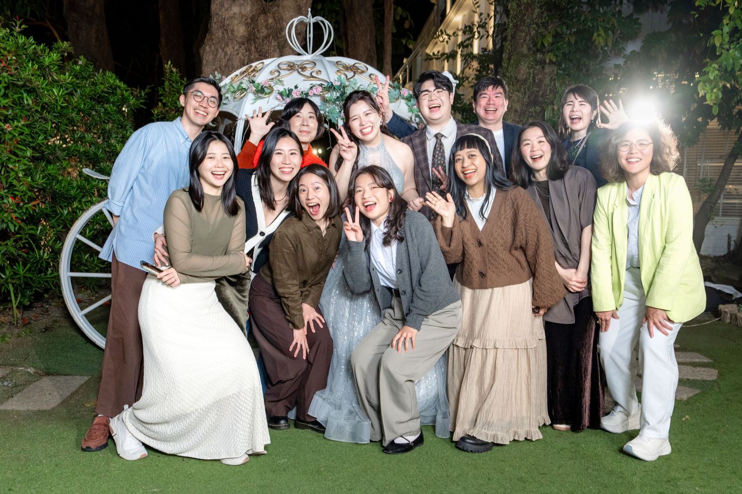 Group photo of people gathered outdoors in front of a decorative carriage wheel wearing casual and formal attire.