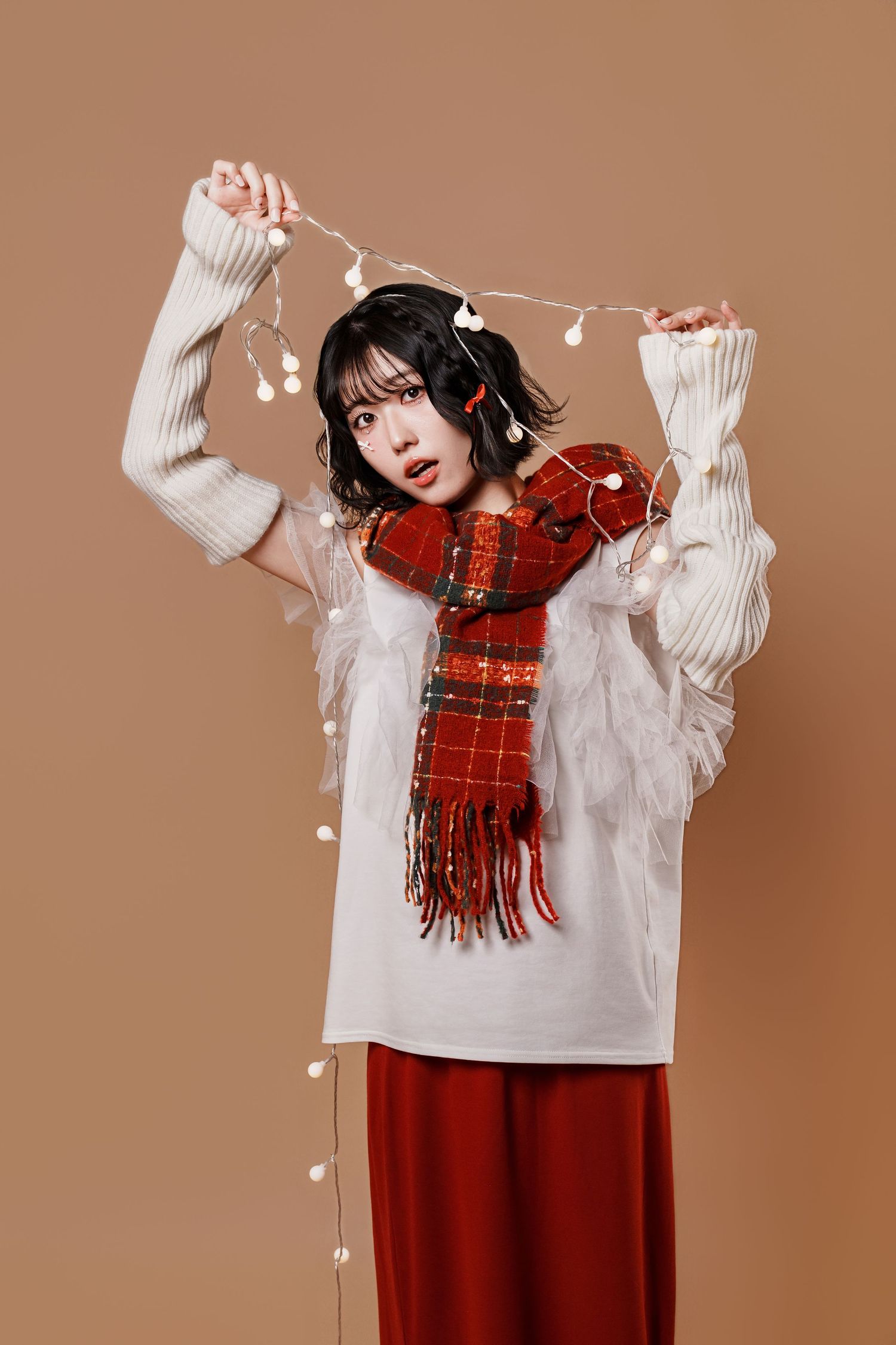 A person in a white sweater and red skirt poses playfully with a red plaid scarf against a brown backdrop.
