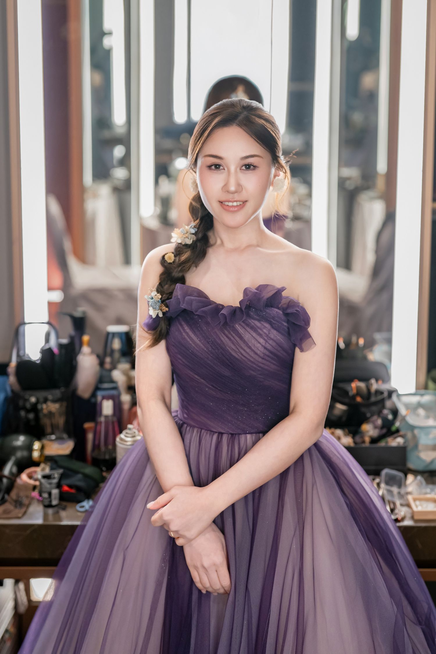 A person poses in a flowing purple tulle gown with ruched bodice in an elegant indoor setting.