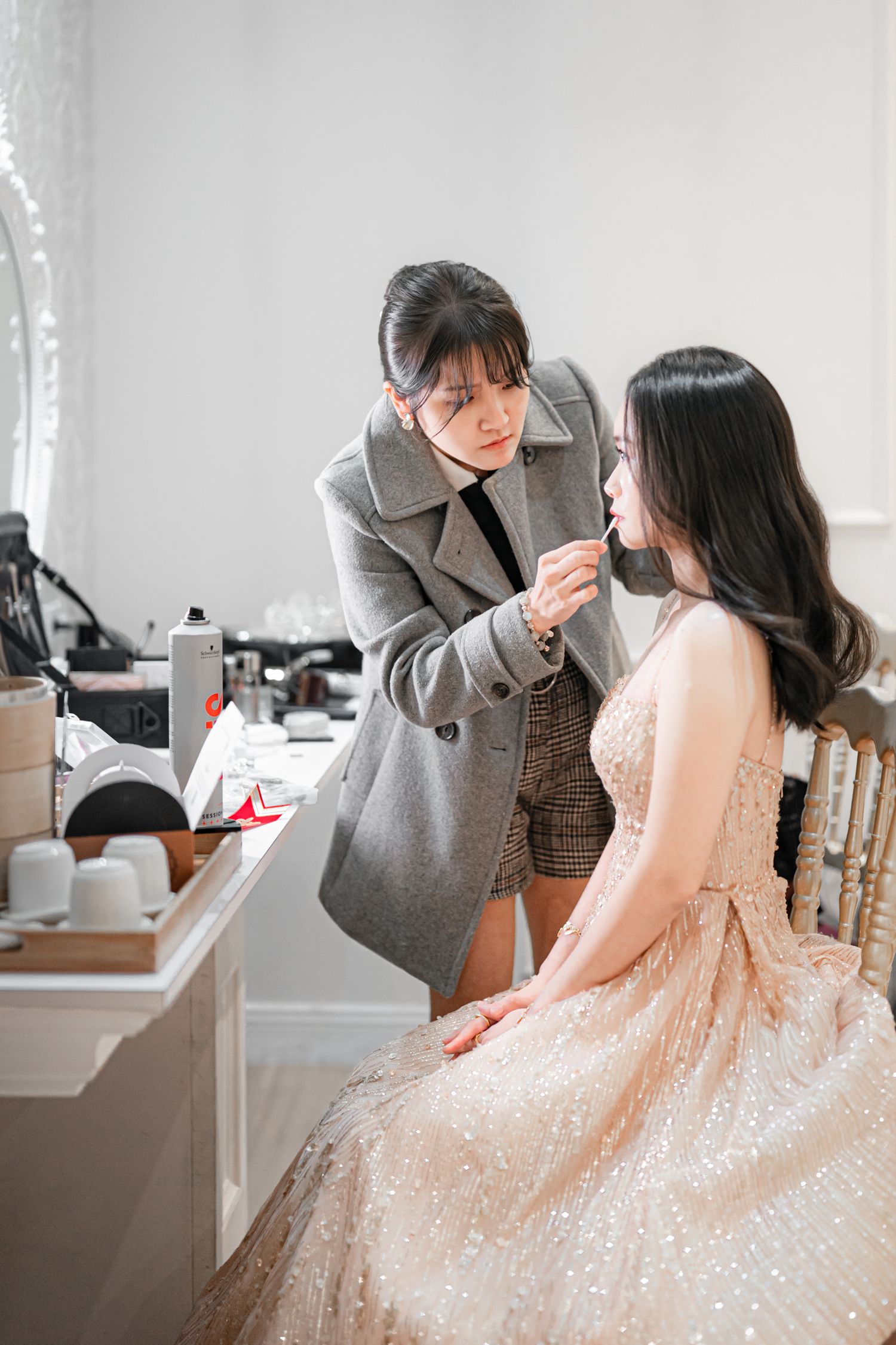 A makeup artist applies cosmetics to a model wearing a sparkly pink dress in a bridal preparation photoshoot series.
