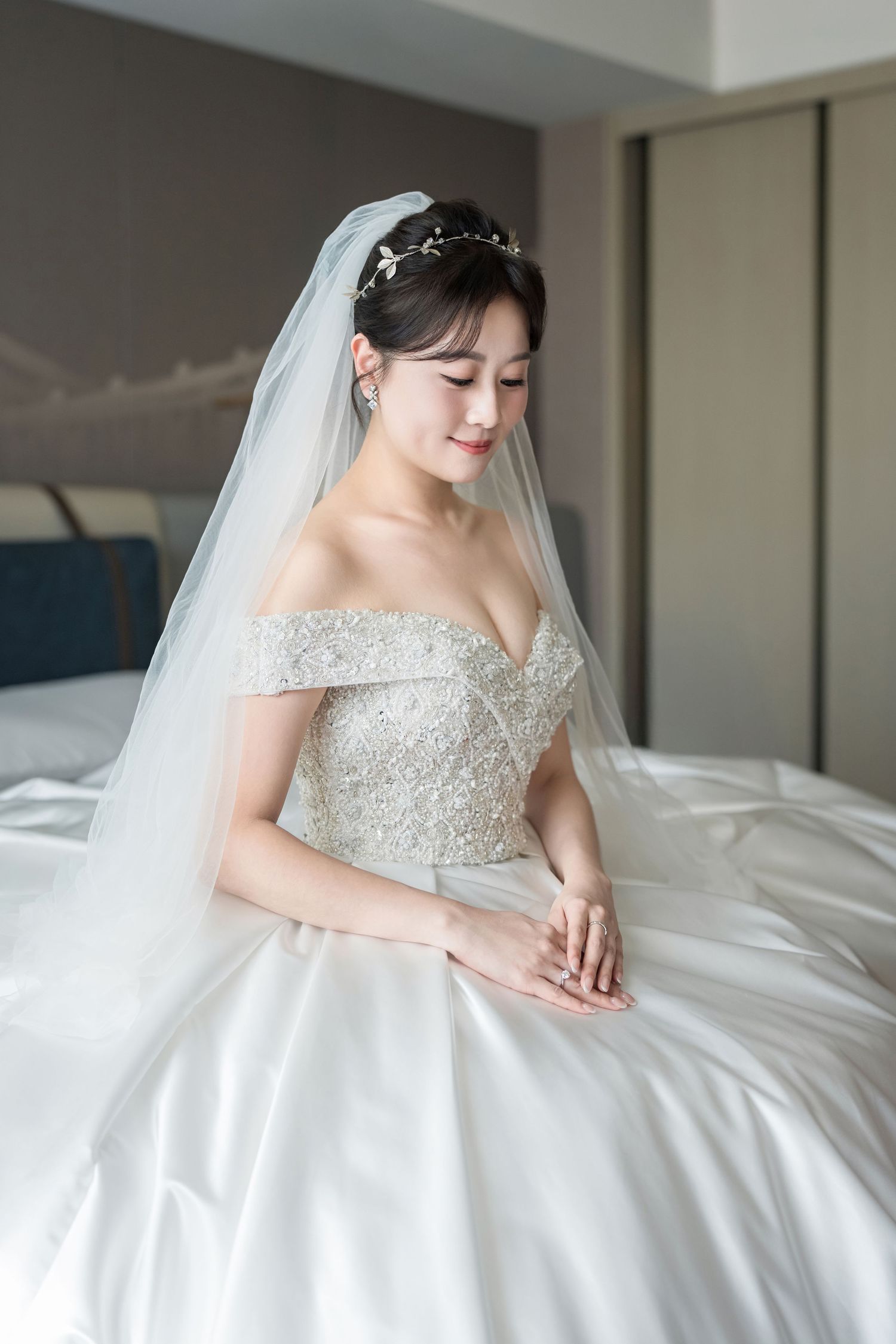 A bride in a white beaded wedding gown and veil sits gracefully on white bedding in an elegant bridal portrait series.