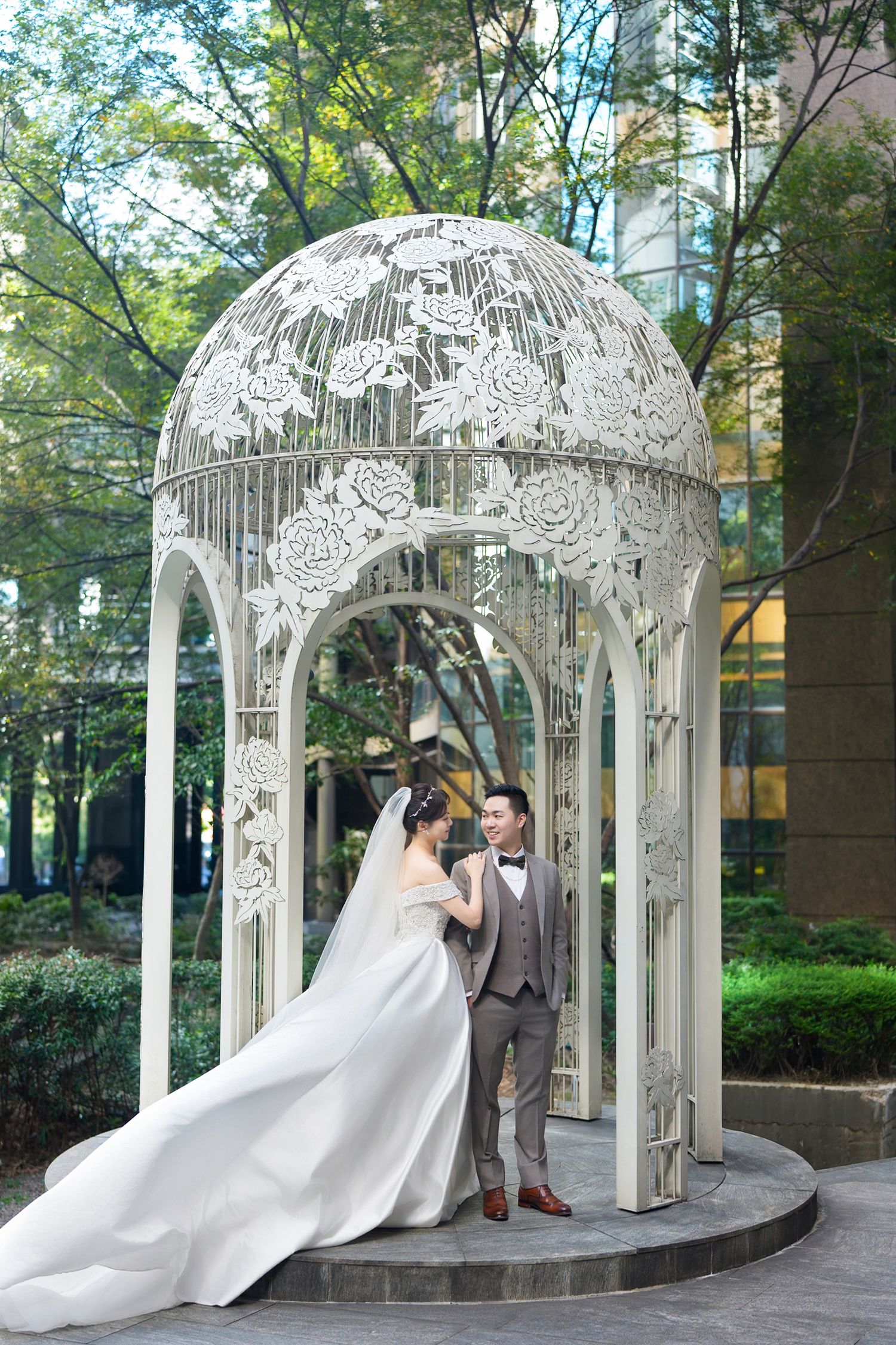 A wedding ceremony takes place under an ornate white metal gazebo with floral designs in a garden setting.