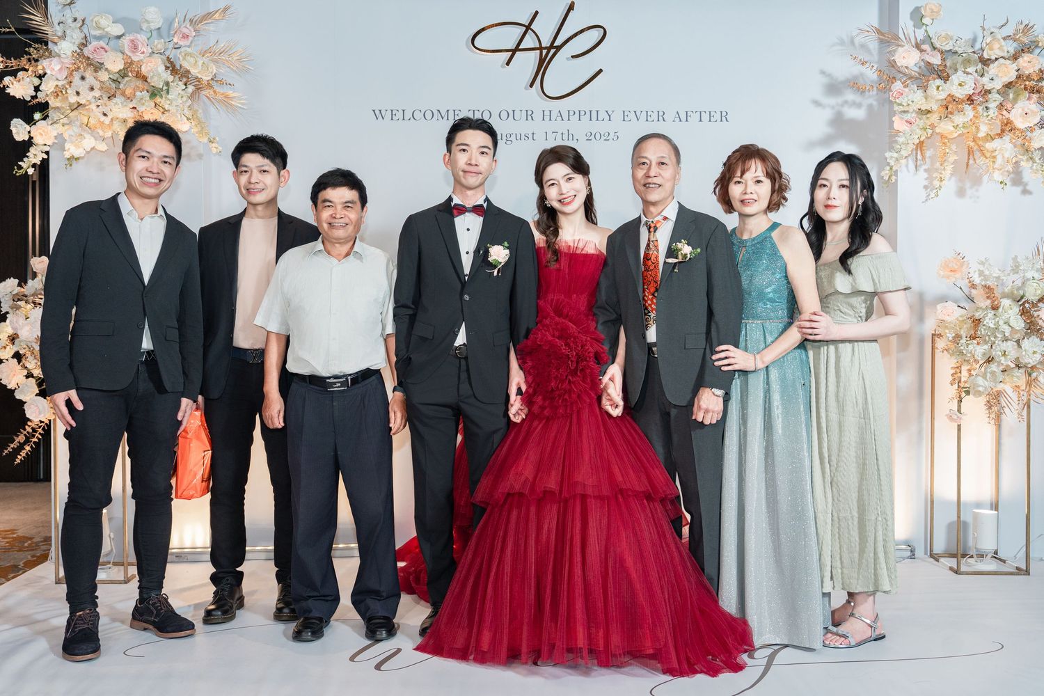 Wedding celebration group photo with bride in red gown and guests posing in front of floral backdrop.