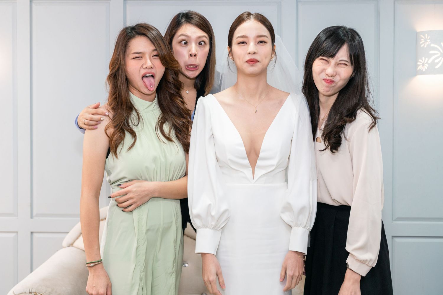 A fun group of four friends in casual attire pose together against a white wall, with one making a playful expression.