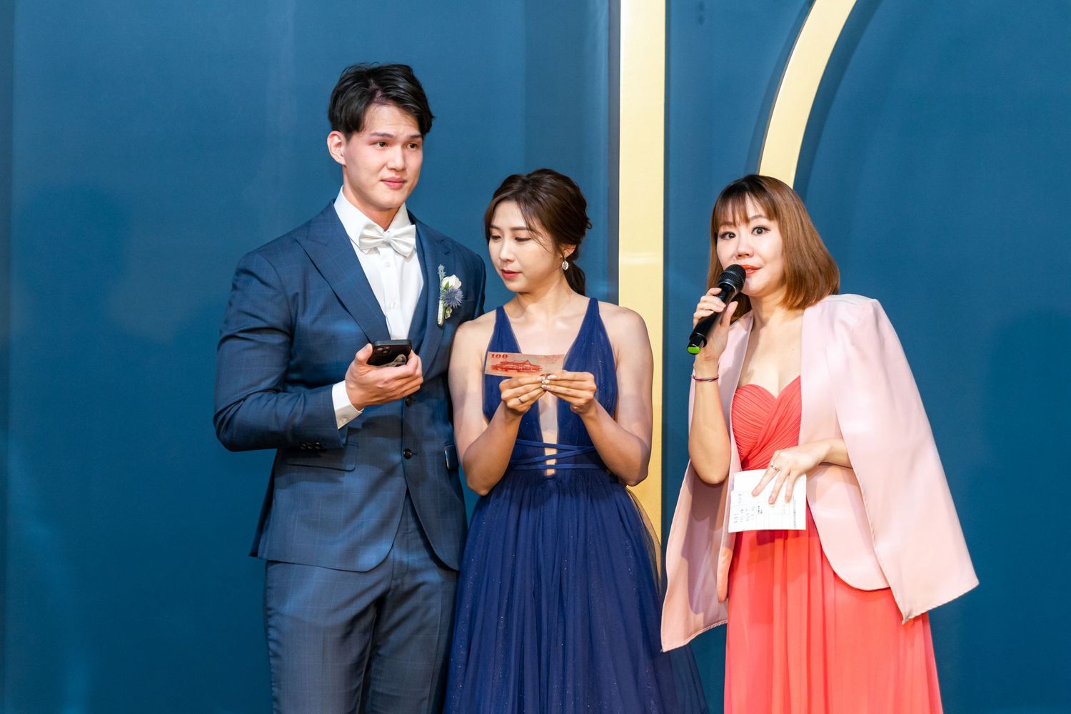 Three people dressed in formal attire stand together on stage sharing a speaking moment against a blue backdrop.