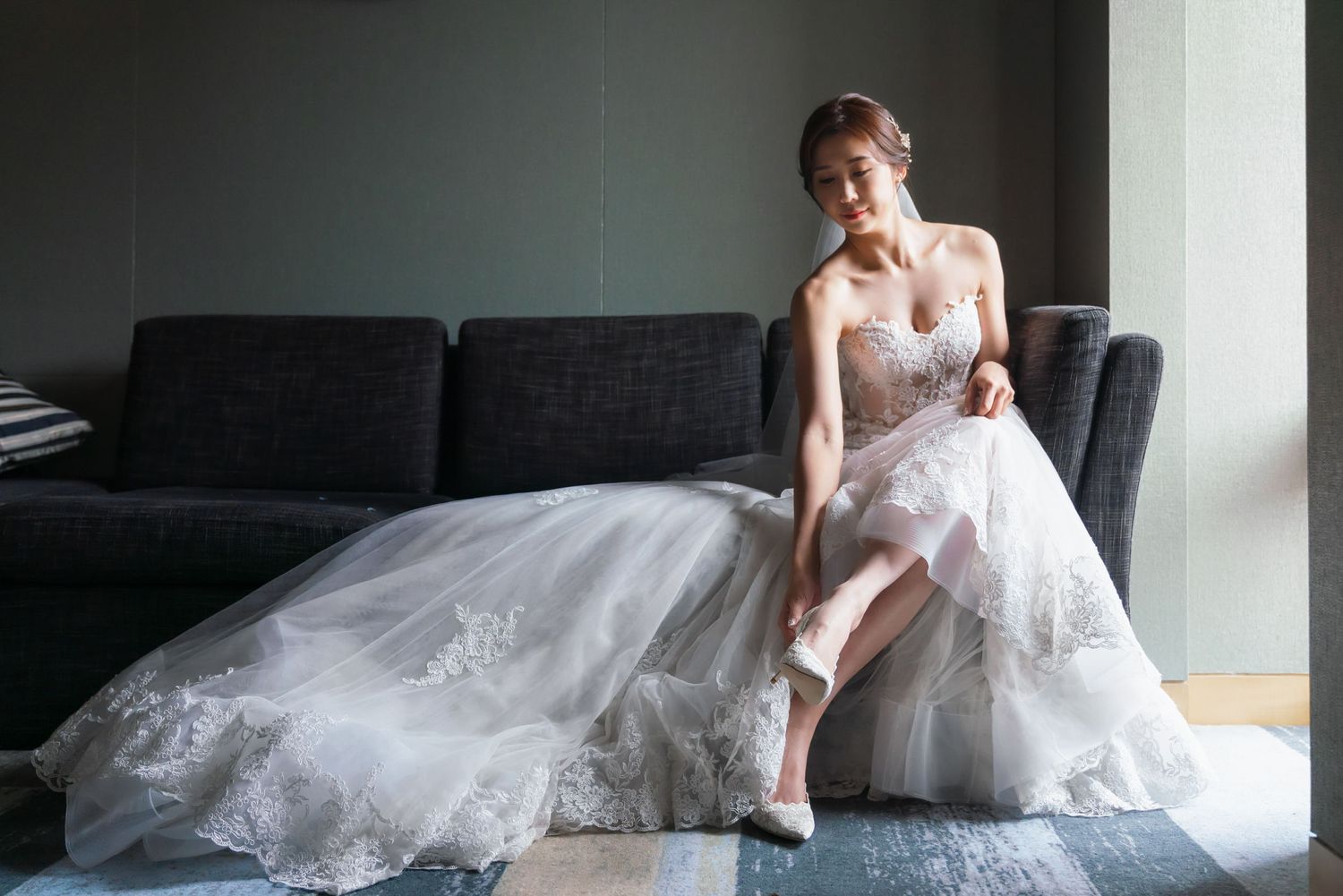 A person in a flowing white wedding dress sits elegantly on a gray sofa while adjusting their shoe.