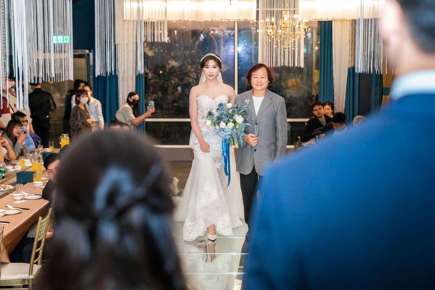 A formal wedding ceremony takes place in an elegant venue with glass walls and crystal chandeliers overhead.