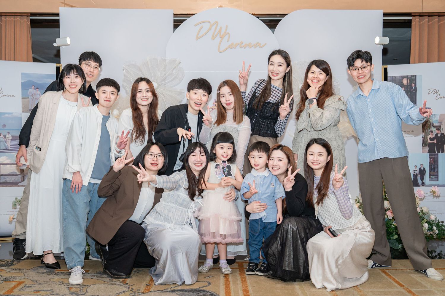 Group of friends posing together at an elegant event with a decorative backdrop, making peace signs and smiling.