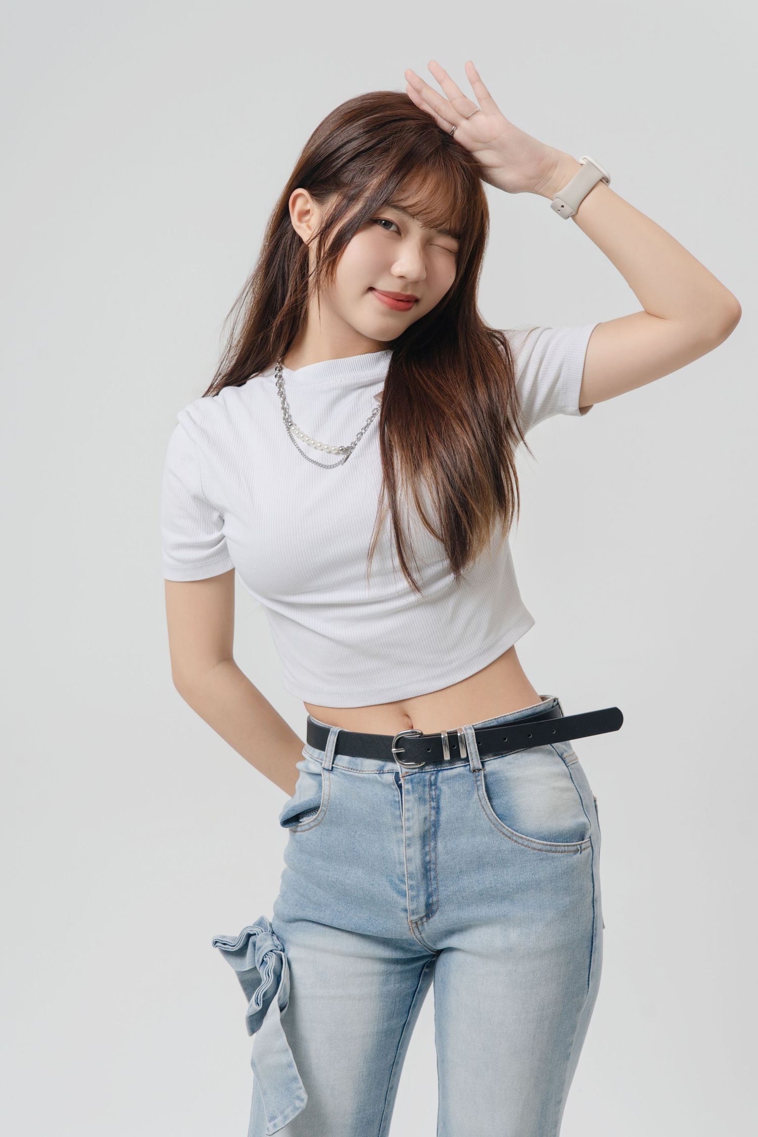 Young woman in white crop top and light jeans poses casually against grey background with hand raised.
