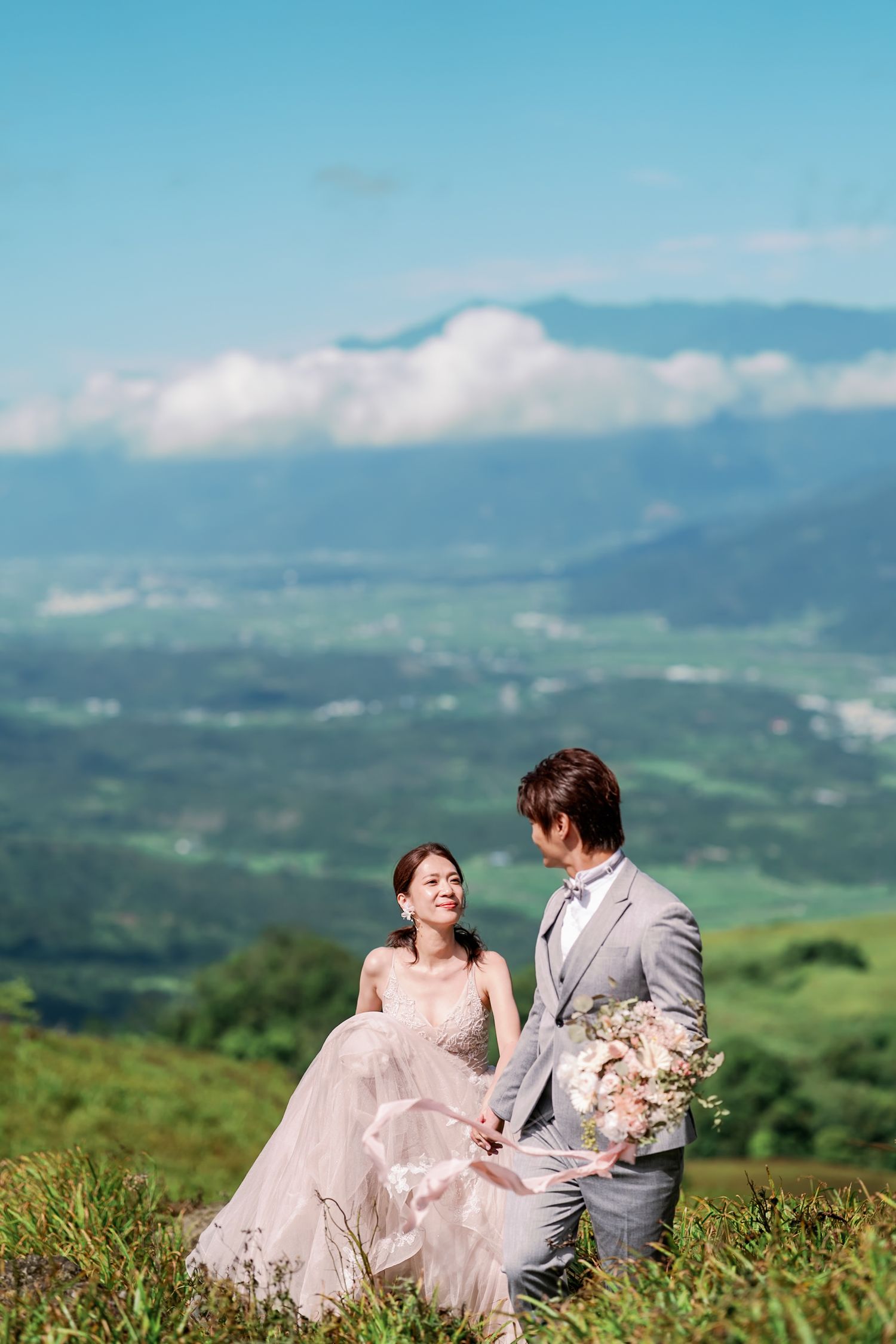 A couple in wedding attire sits together on a grassy hilltop overlooking a scenic mountain landscape on a sunny day.