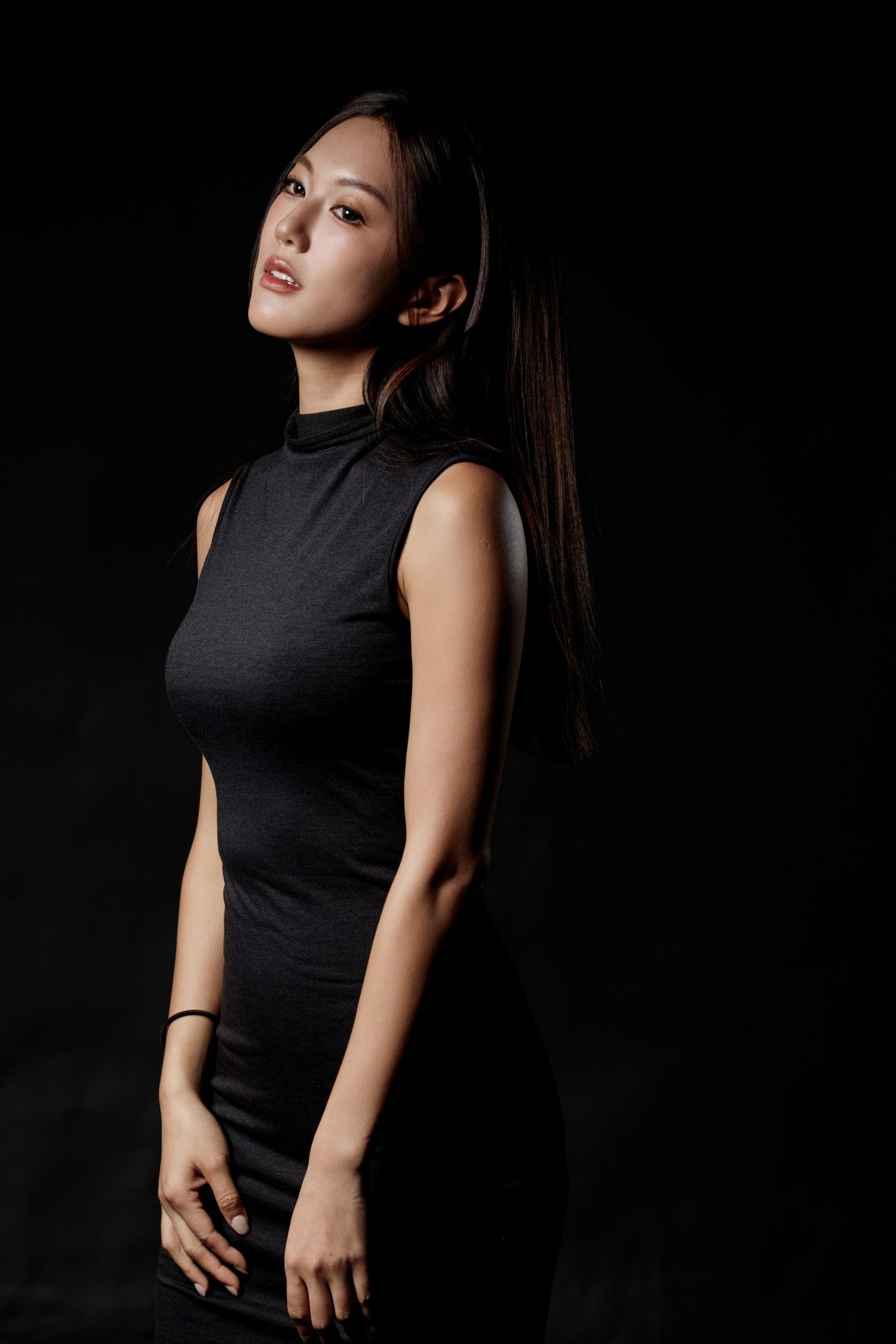 Young Asian woman in sleek black sleeveless dress posing confidently against dark studio background.