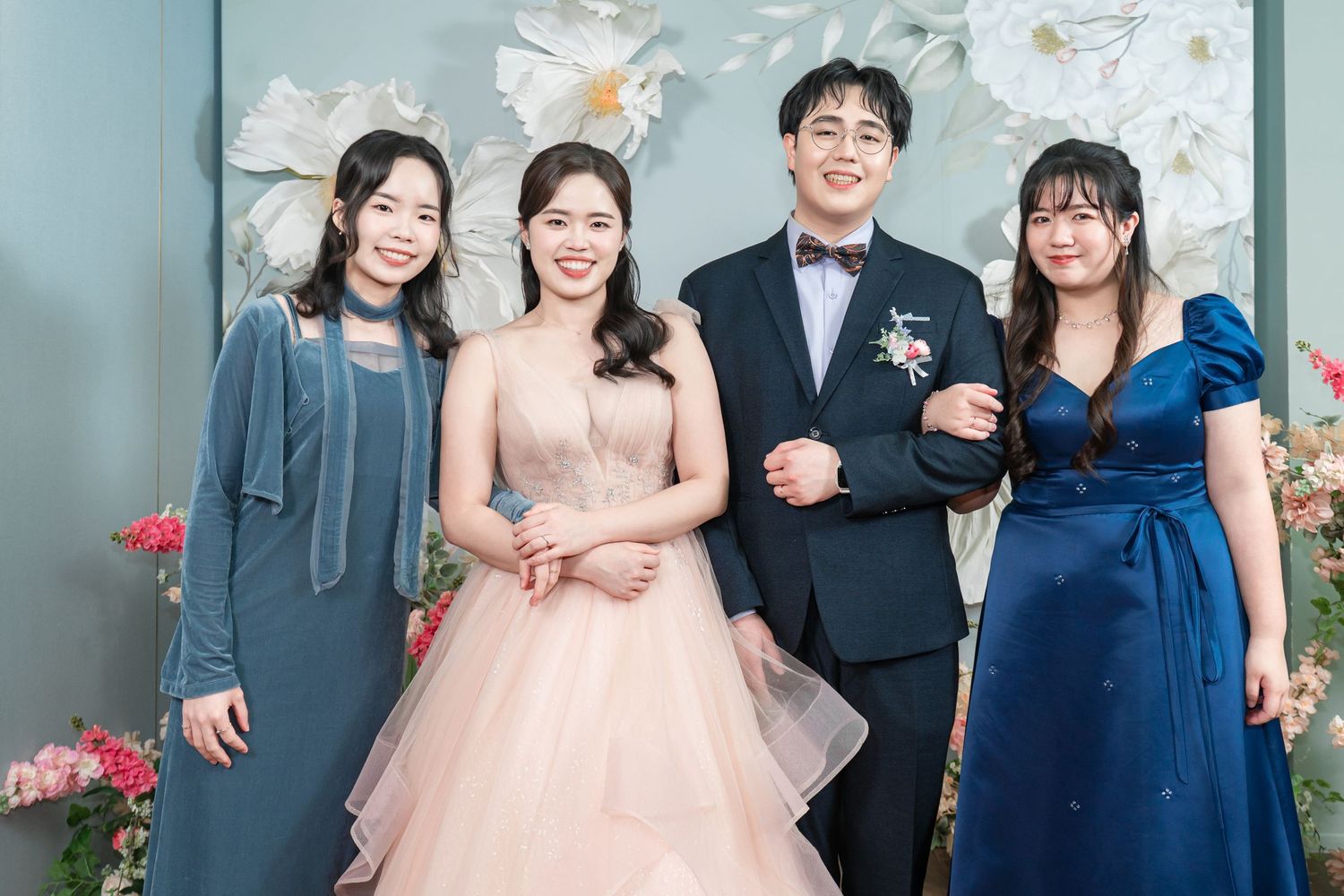 Wedding party members pose together in formal attire with floral decorations in the background.