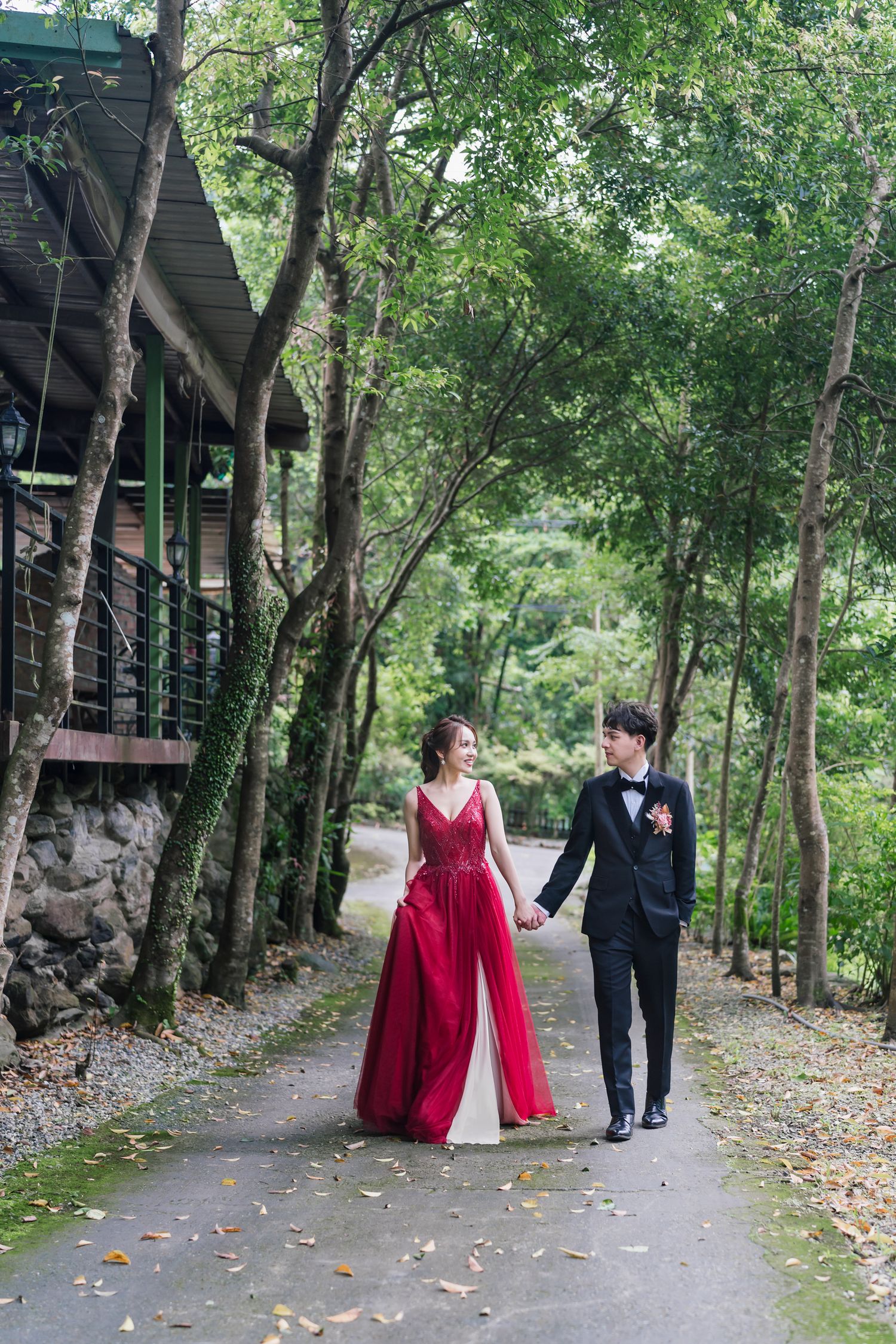 Elegant couple strolling hand in hand through wooded path in formal attire.