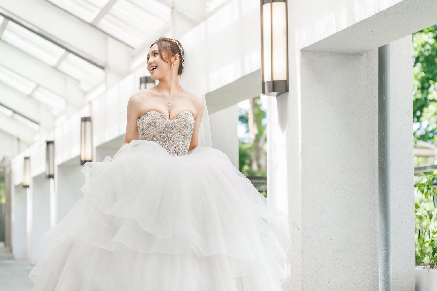Bride in strapless beaded gown stands in sunlit corridor with modern architectural details.