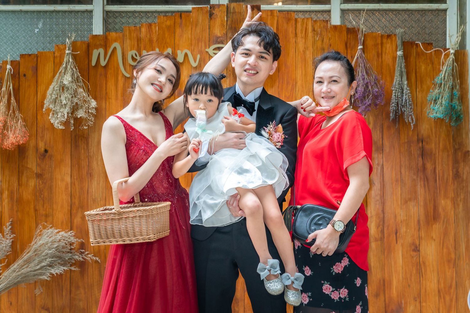 Small group poses joyfully with young child against rustic wooden wall with dried flowers.