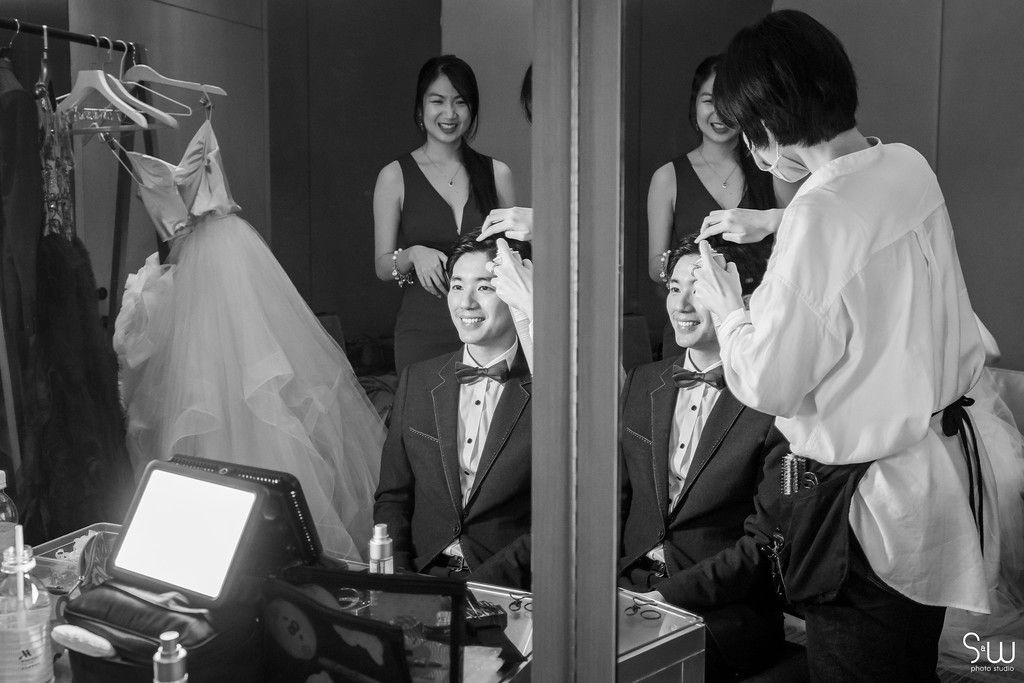 Behind-the-scenes black and white photo of wedding preparation with mirrors and formal attire.