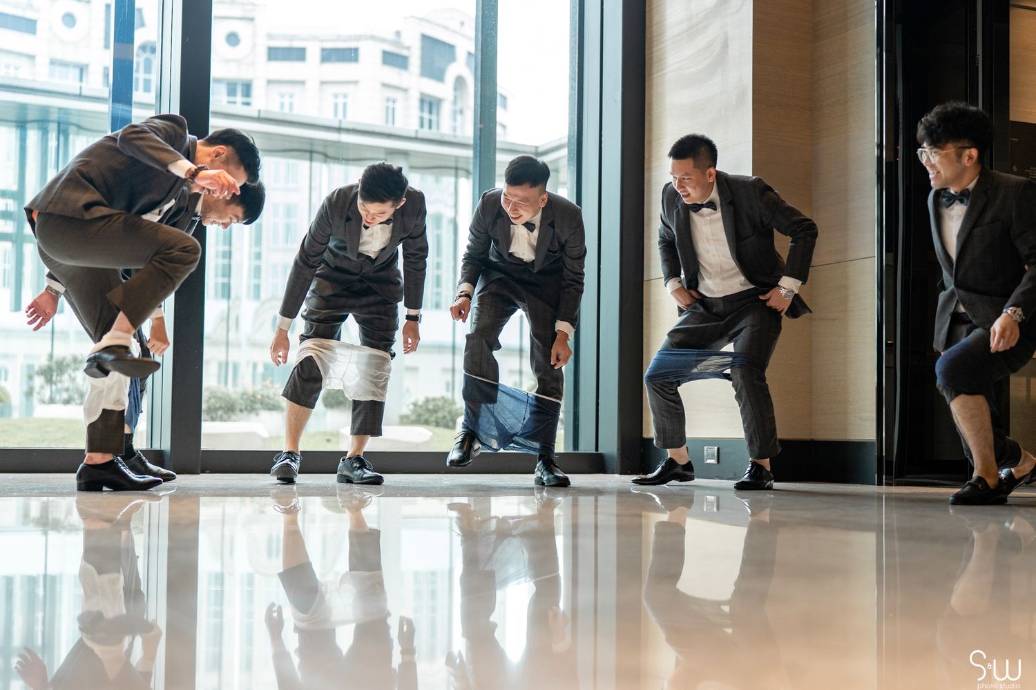 Groomsmen in suits strike playful poses against large windows in a modern building hallway.