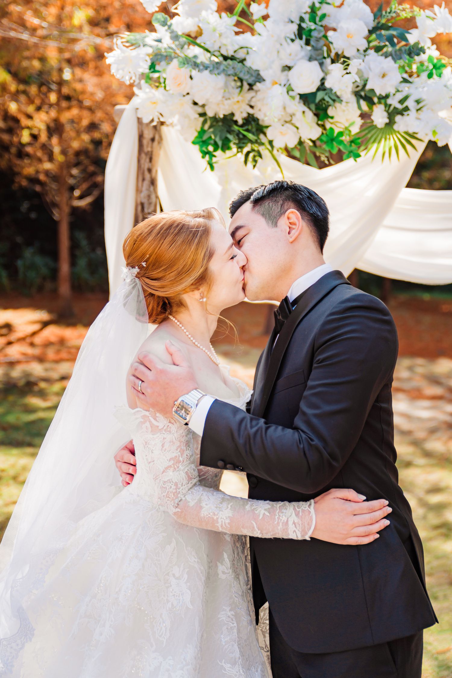 Newlyweds share romantic moment under white floral arch with autumn foliage backdrop.