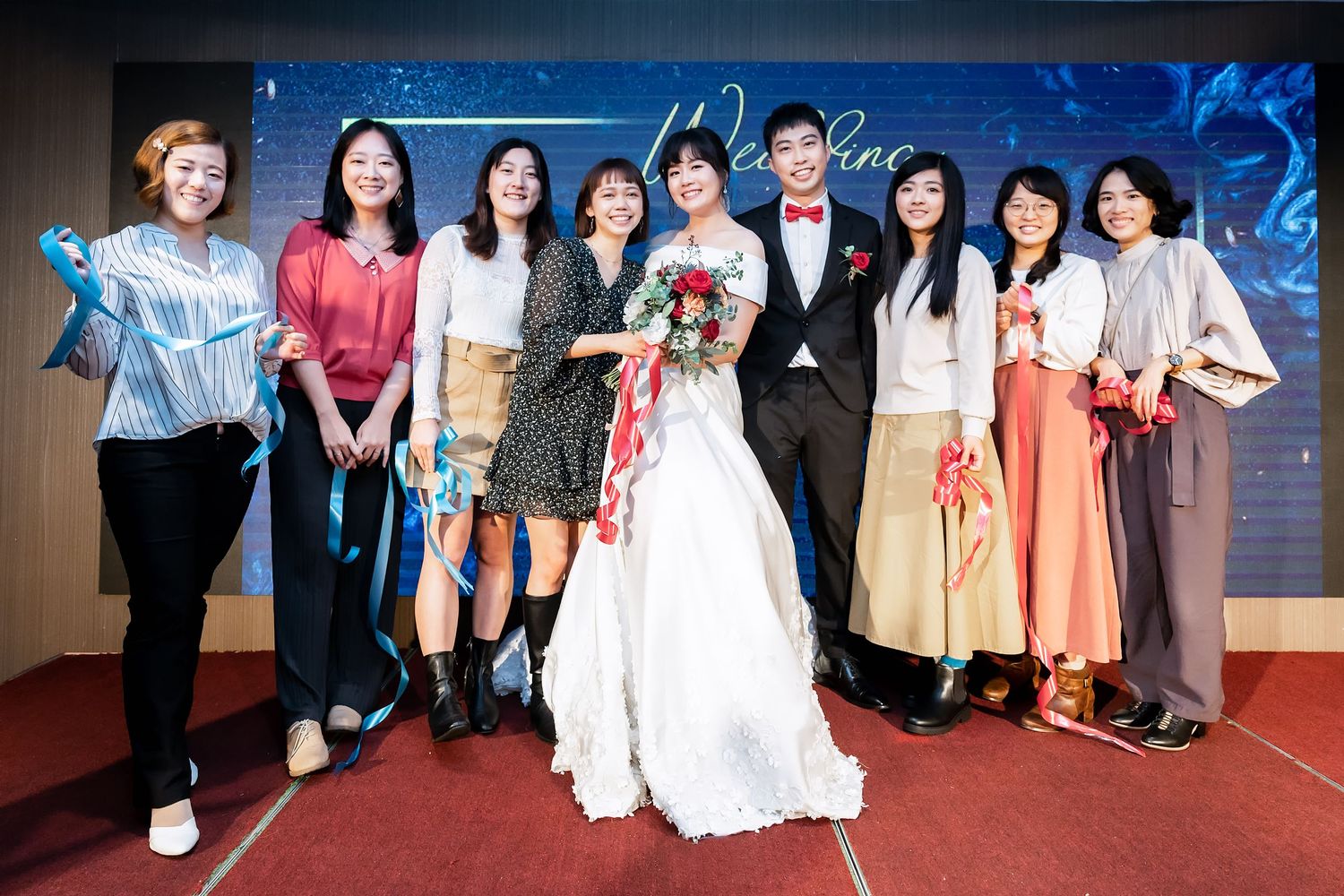 Wedding party poses for formal group photo on red carpet with decorative blue backdrop.