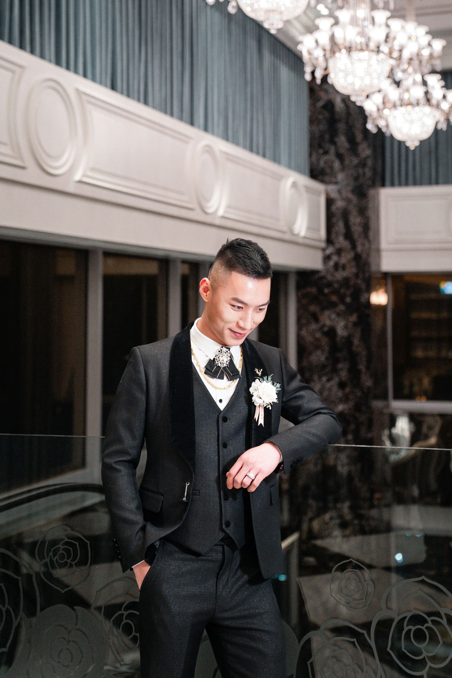 Elegant black three-piece suit with bow tie and boutonniere in luxurious venue with crystal chandelier and ornate decor.