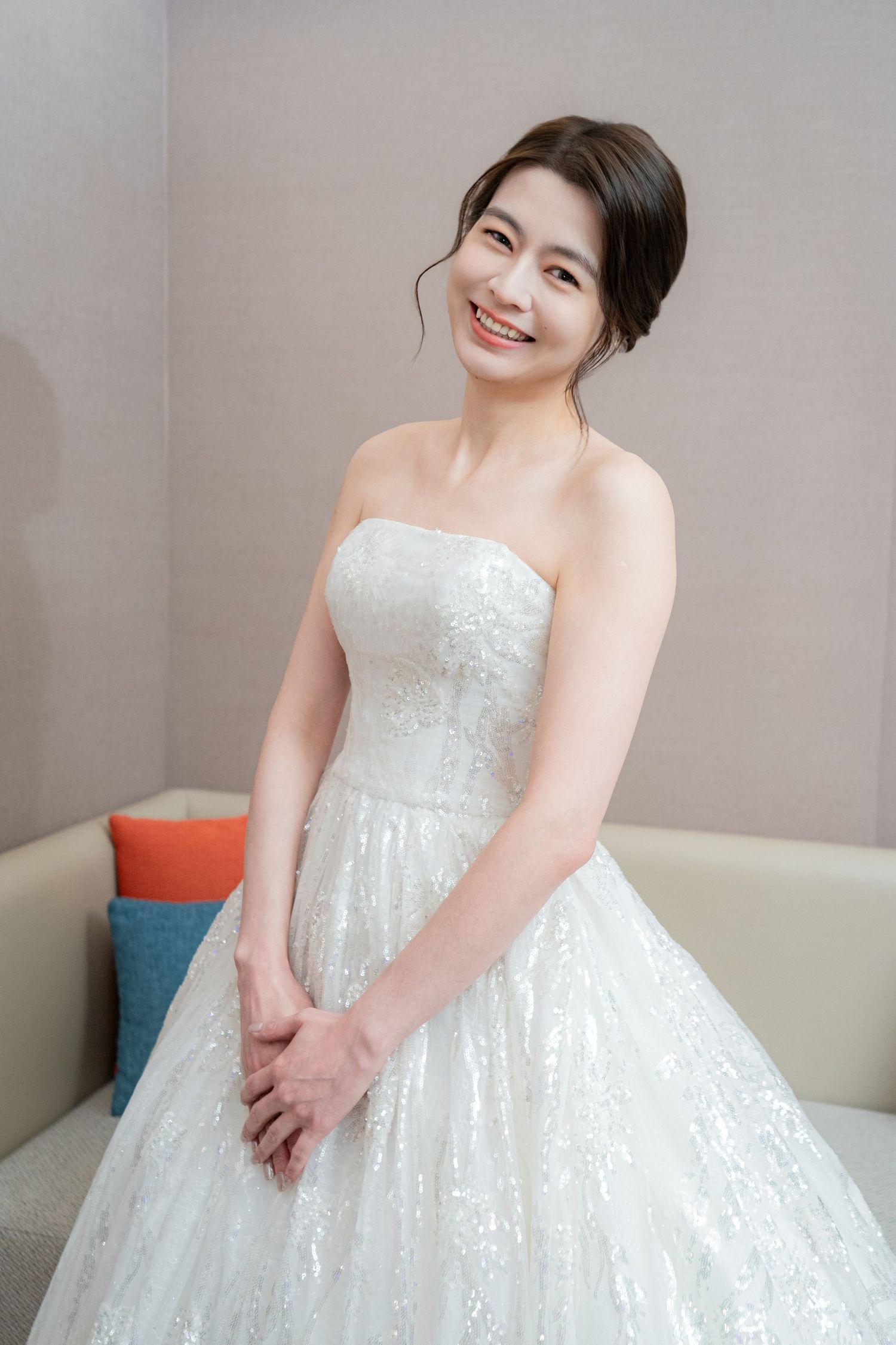 A bride in a white strapless sequined ball gown smiles while sitting on a gray couch with colorful throw pillows.