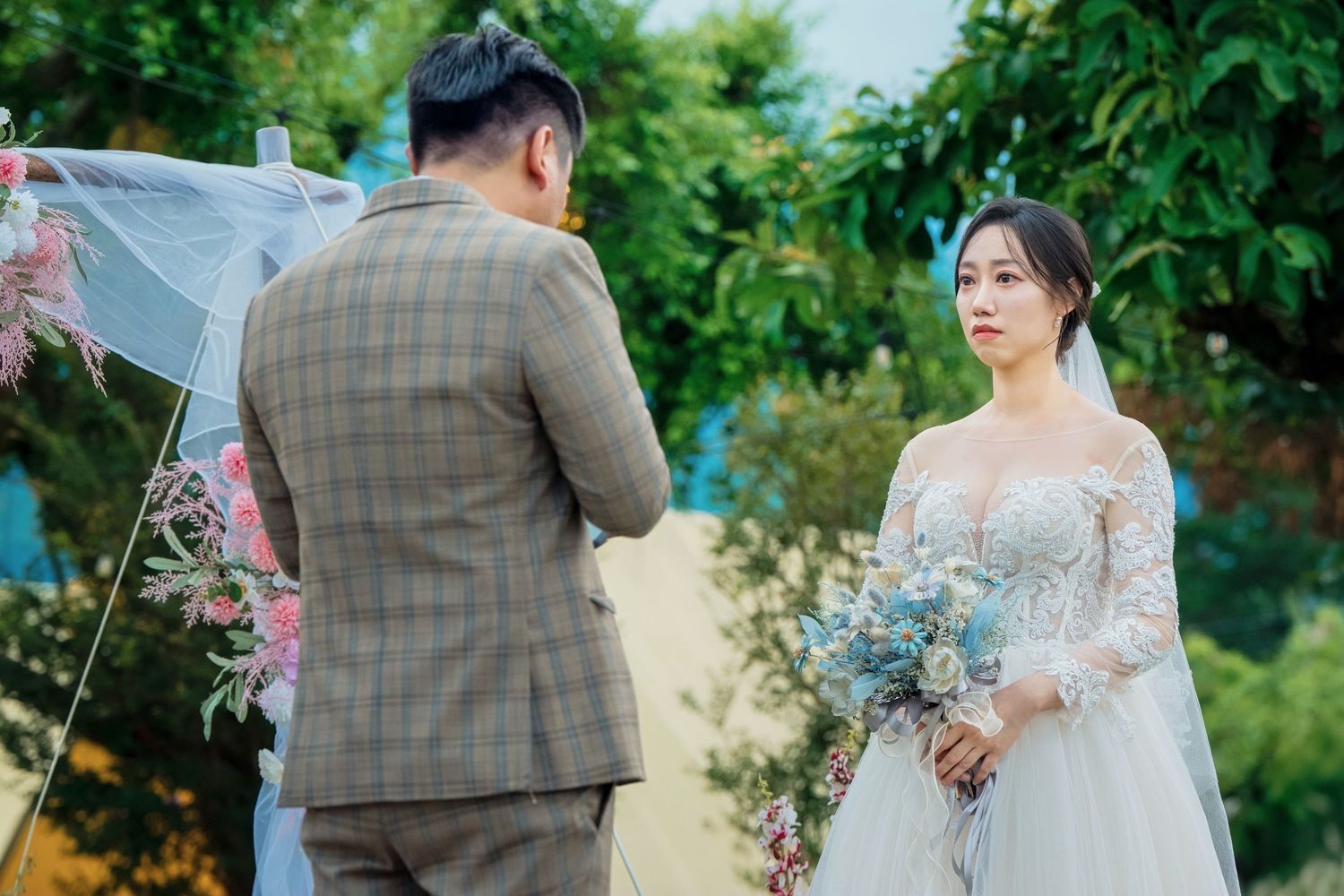 Newlywed couple walking through a garden path with green foliage and trees in the background.