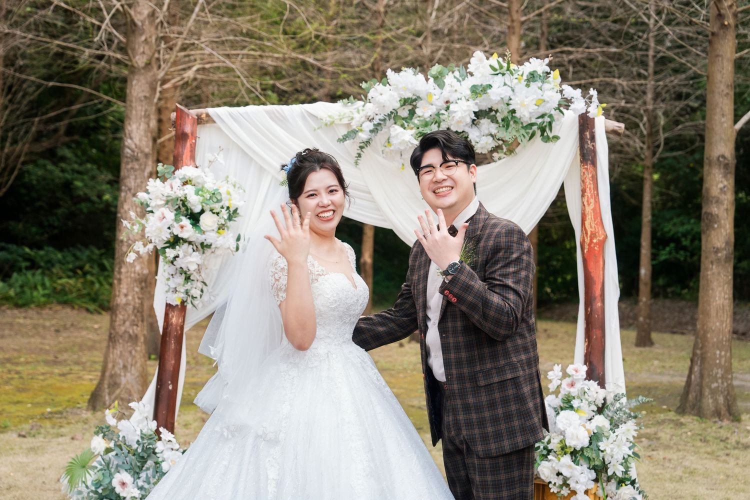 A wedding couple shares joyful moments under a white floral arch in an outdoor garden setting.
