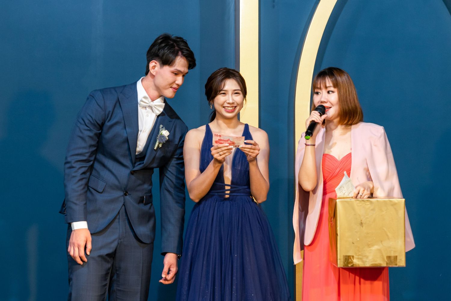 Three friends dressed in formal attire stand together against a blue wall, smiling and interacting socially.