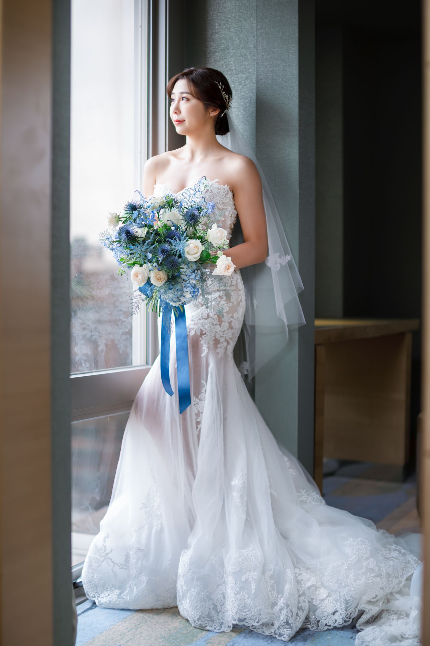 A bride in a white lace wedding gown holds a blue and white bouquet while standing near a window.