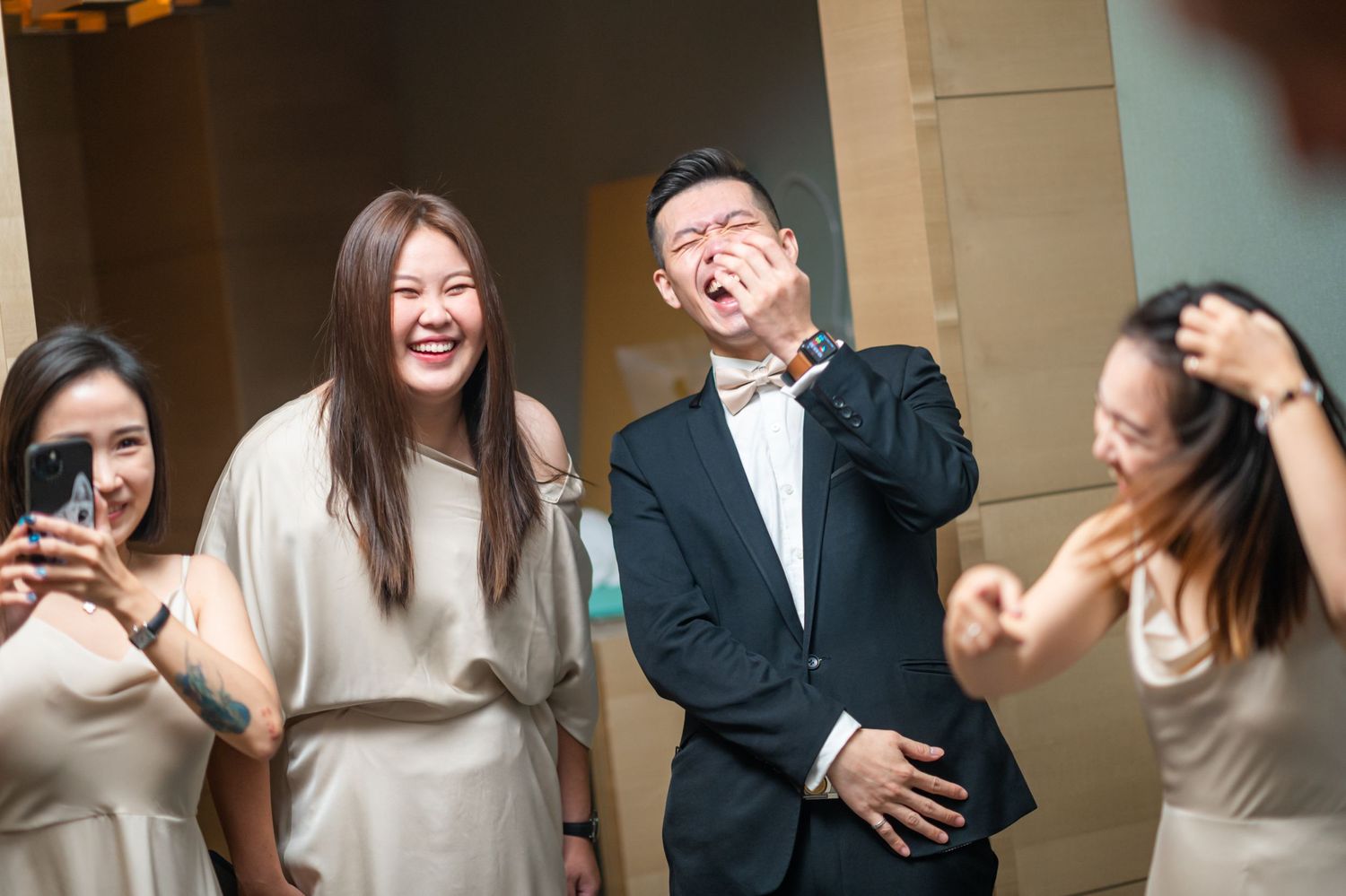 A group of friends in formal and casual attire laugh and pose playfully together at an indoor event.