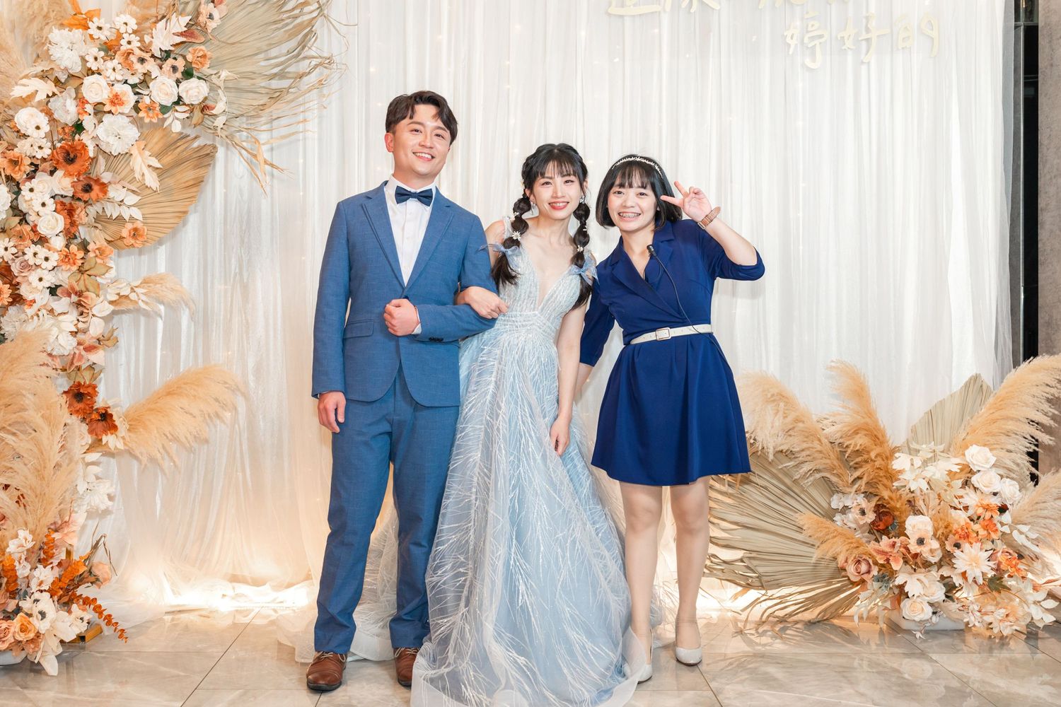 Three people dressed in formal attire pose together in front of a floral backdrop with peach and white flowers.
