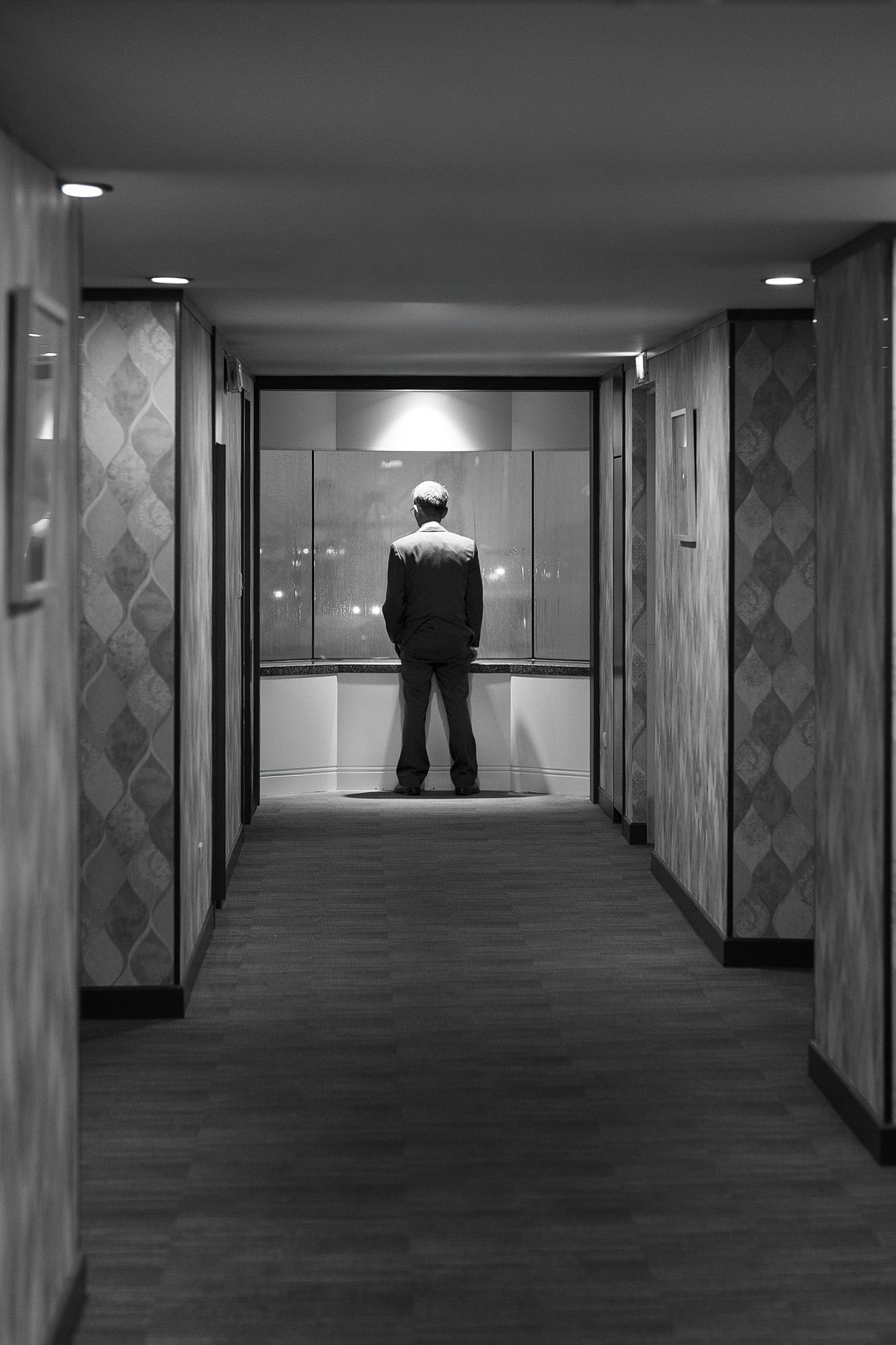 A silhouetted figure stands alone in a dimly lit elevator at the end of a long hallway with patterned walls.