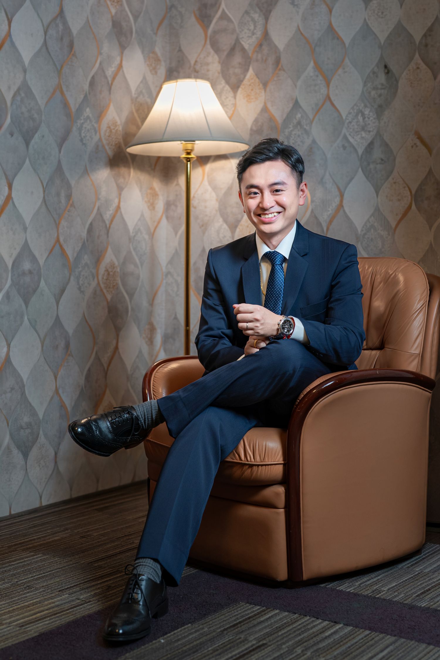 Business professional in navy suit relaxes in brown leather armchair against geometric wallpaper in elegant setting.
