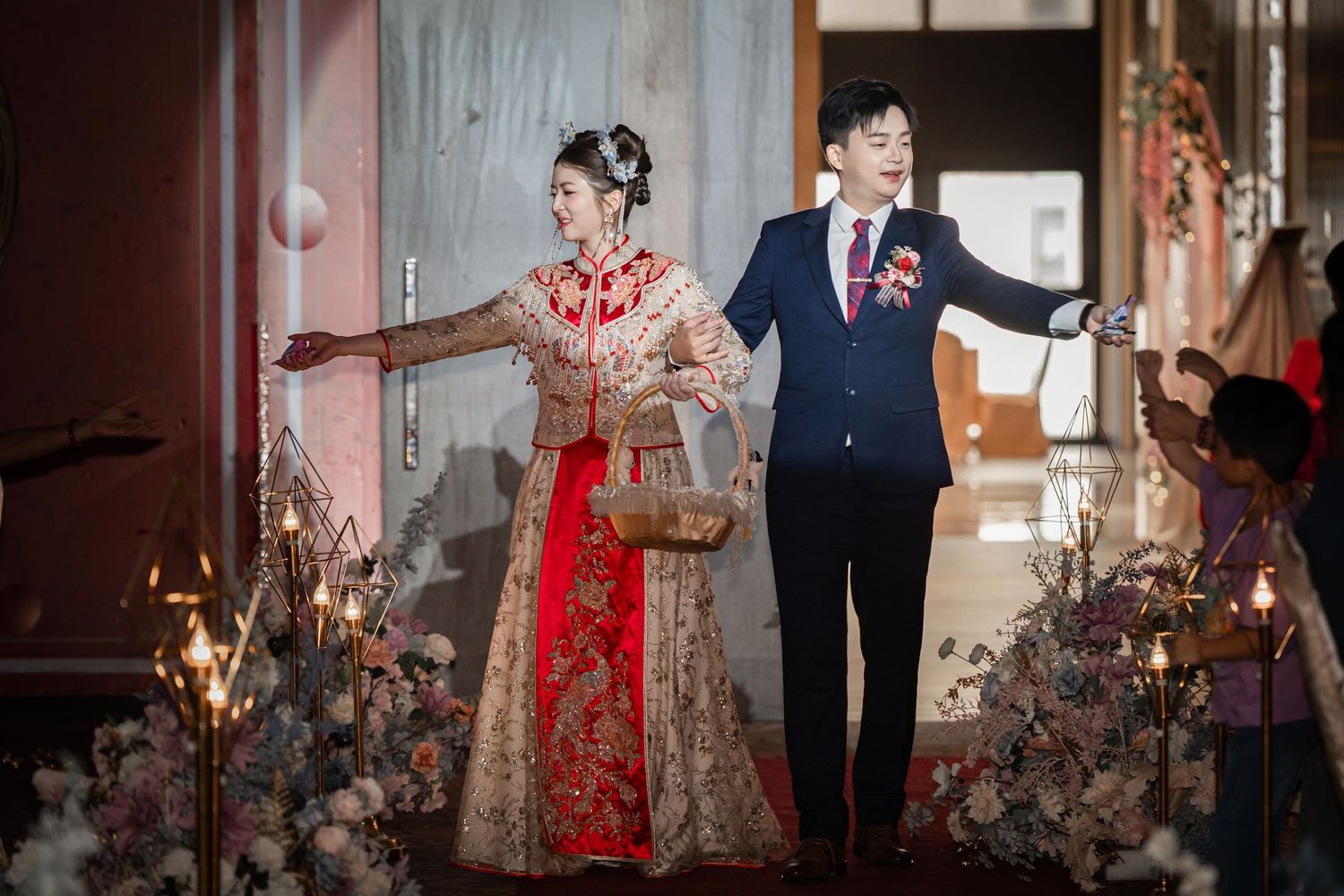 Bride in red and gold embroidered Chinese wedding gown and groom in navy suit walk hand in hand past candles and flowers at a wedding.