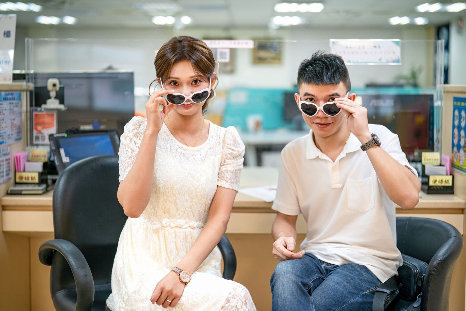 People in white outfits sit at desks in an office setting with bright fluorescent lighting overhead.