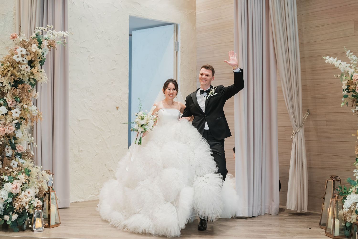 A bride and groom dance together in an elegant ballroom decorated with floral arrangements and soft curtains.
