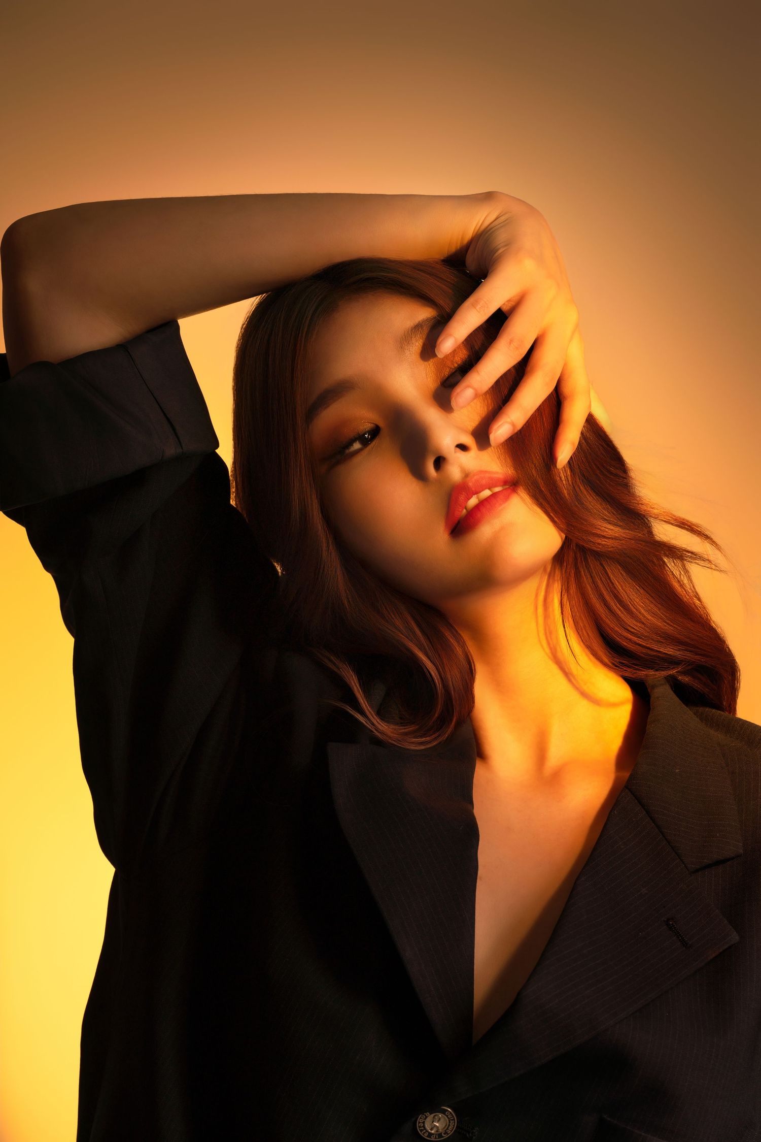 Dramatic portrait in warm golden lighting with artistic pose against yellow background wearing black blazer.