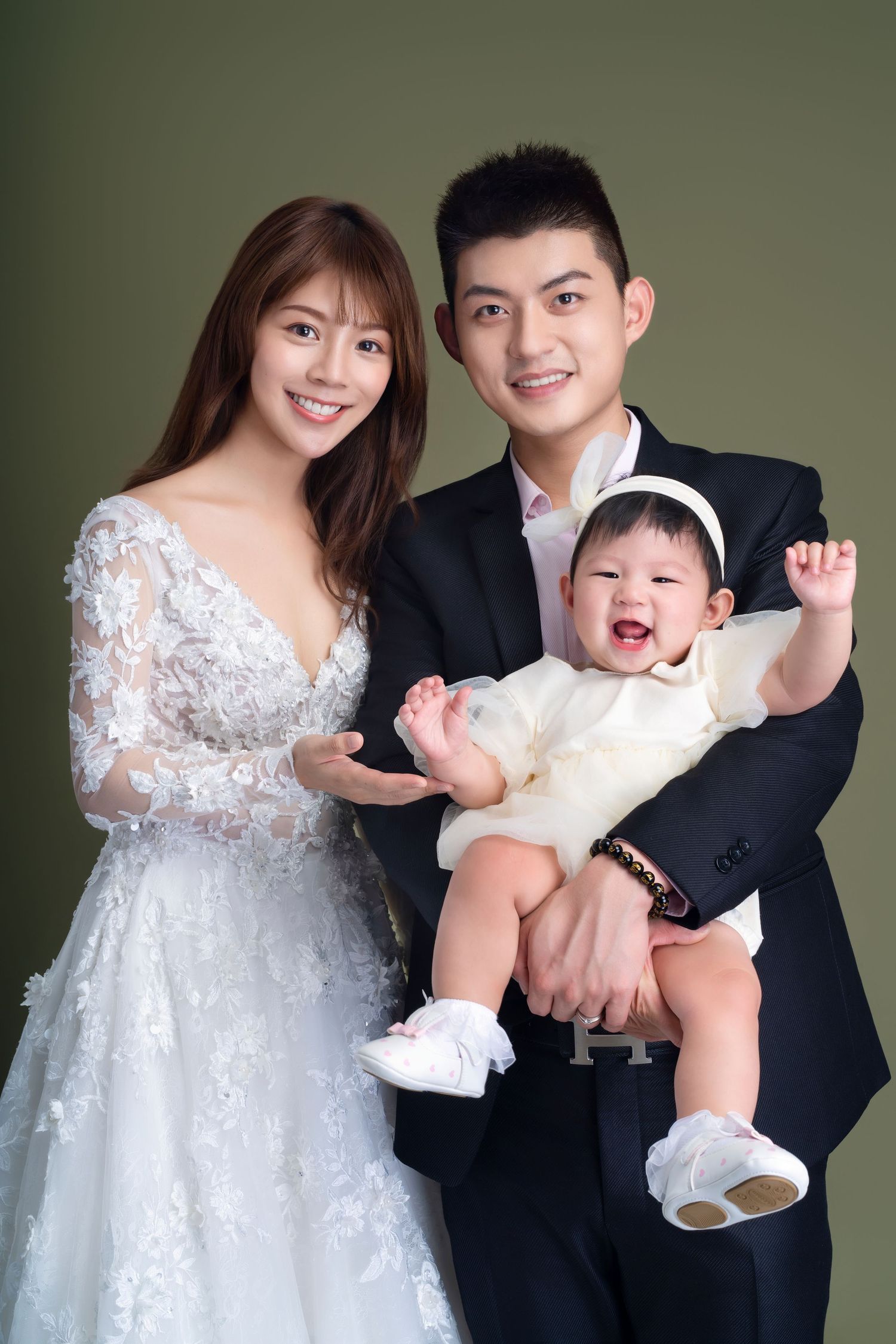 A joyful wedding portrait with a couple in formal attire holding their laughing baby in white against a green backdrop.