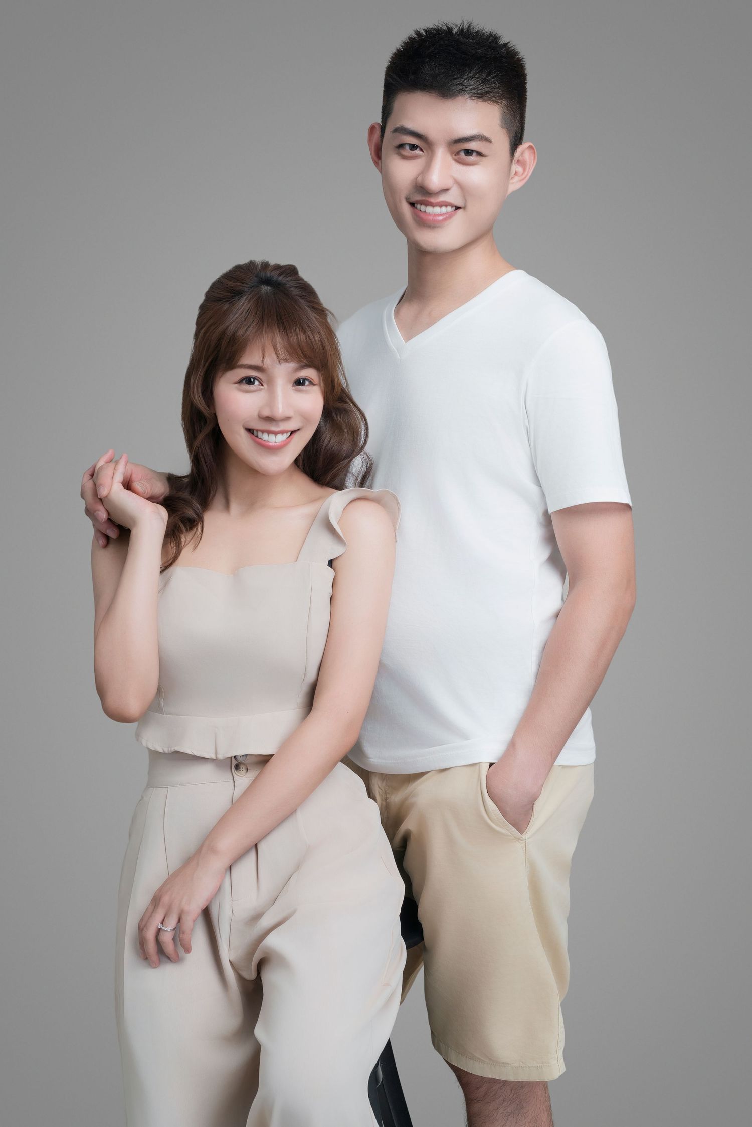 A couple in light colored casual clothing poses together against a gray studio backdrop.