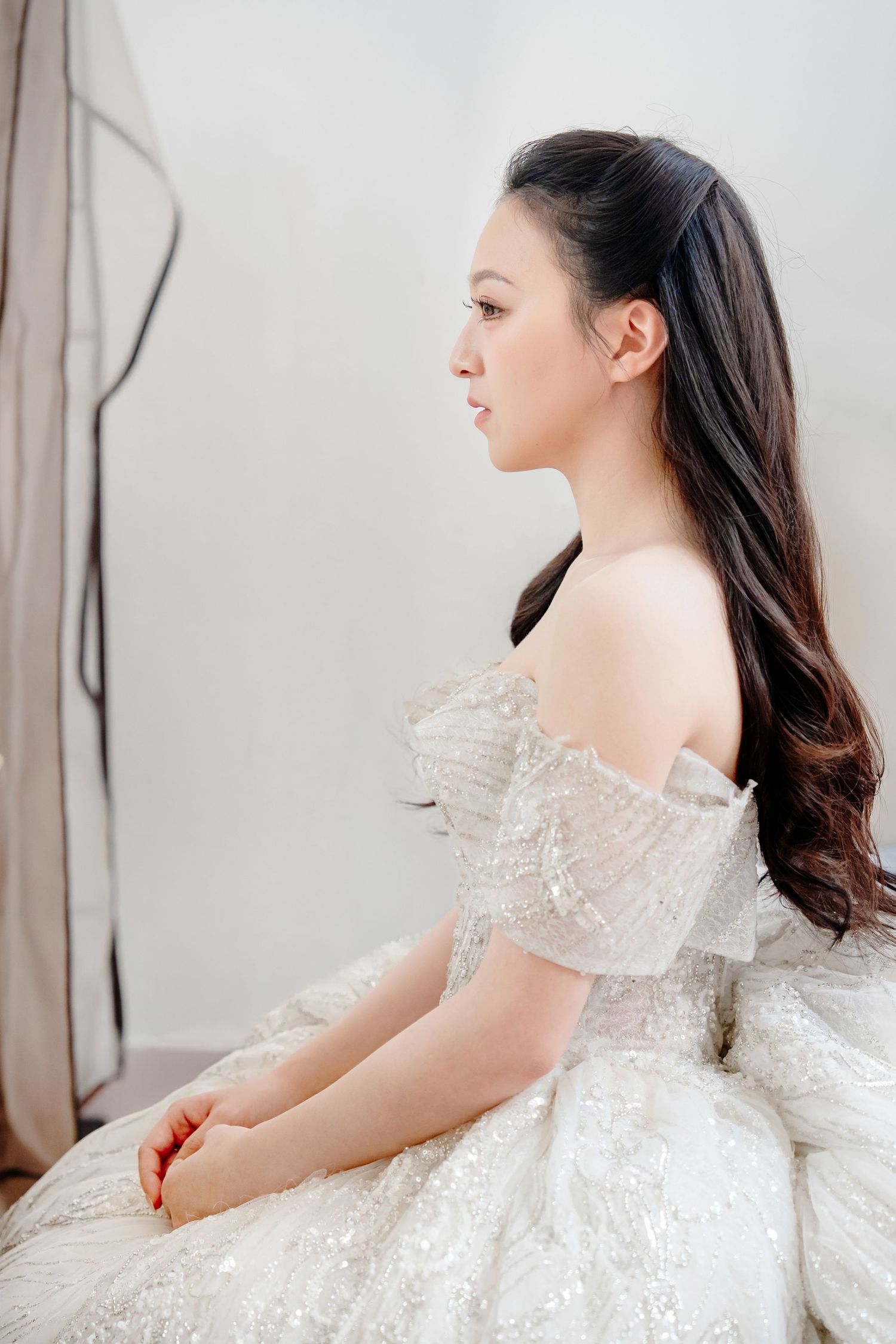 Young Asian woman in elegant off-shoulder white sequined ball gown sits gracefully, hair styled in a long ponytail.