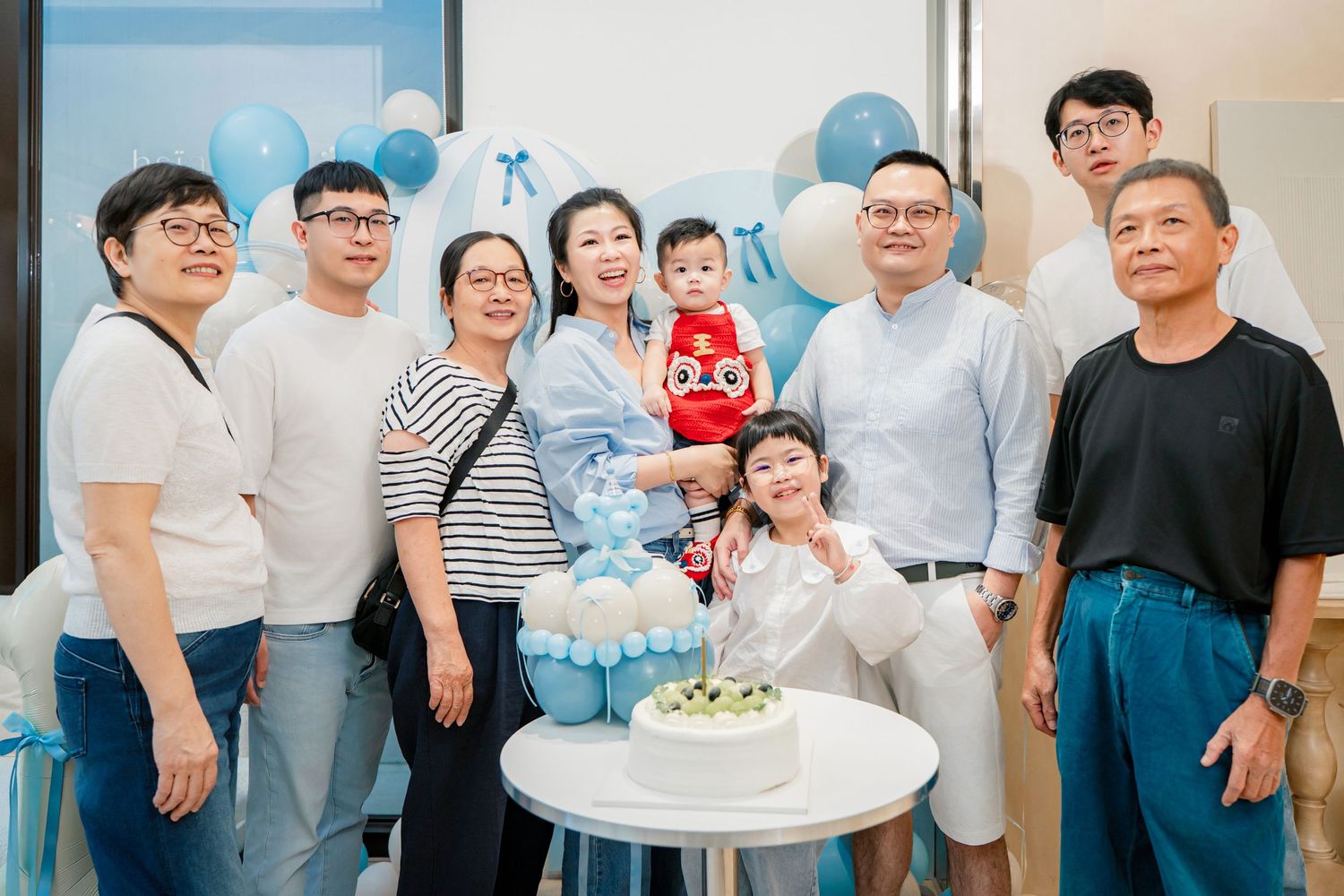 A group of people gather together at a celebration event with blue and white decorations and balloons.