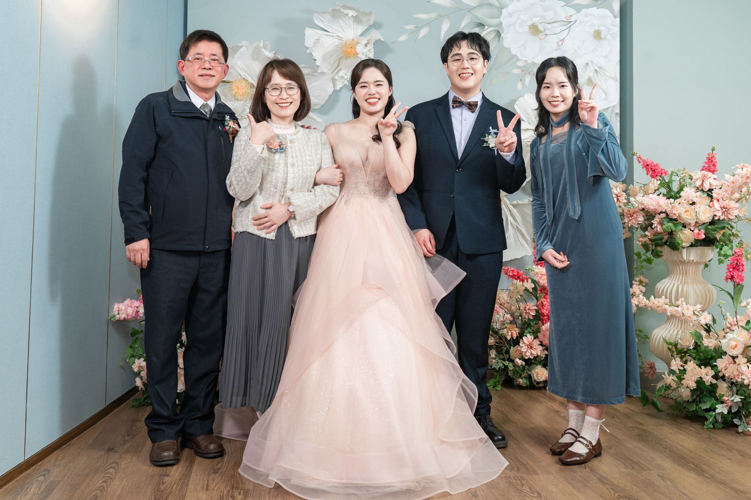 A group poses together in formal attire with pink dresses and dark suits against a blue wall with floral decorations.