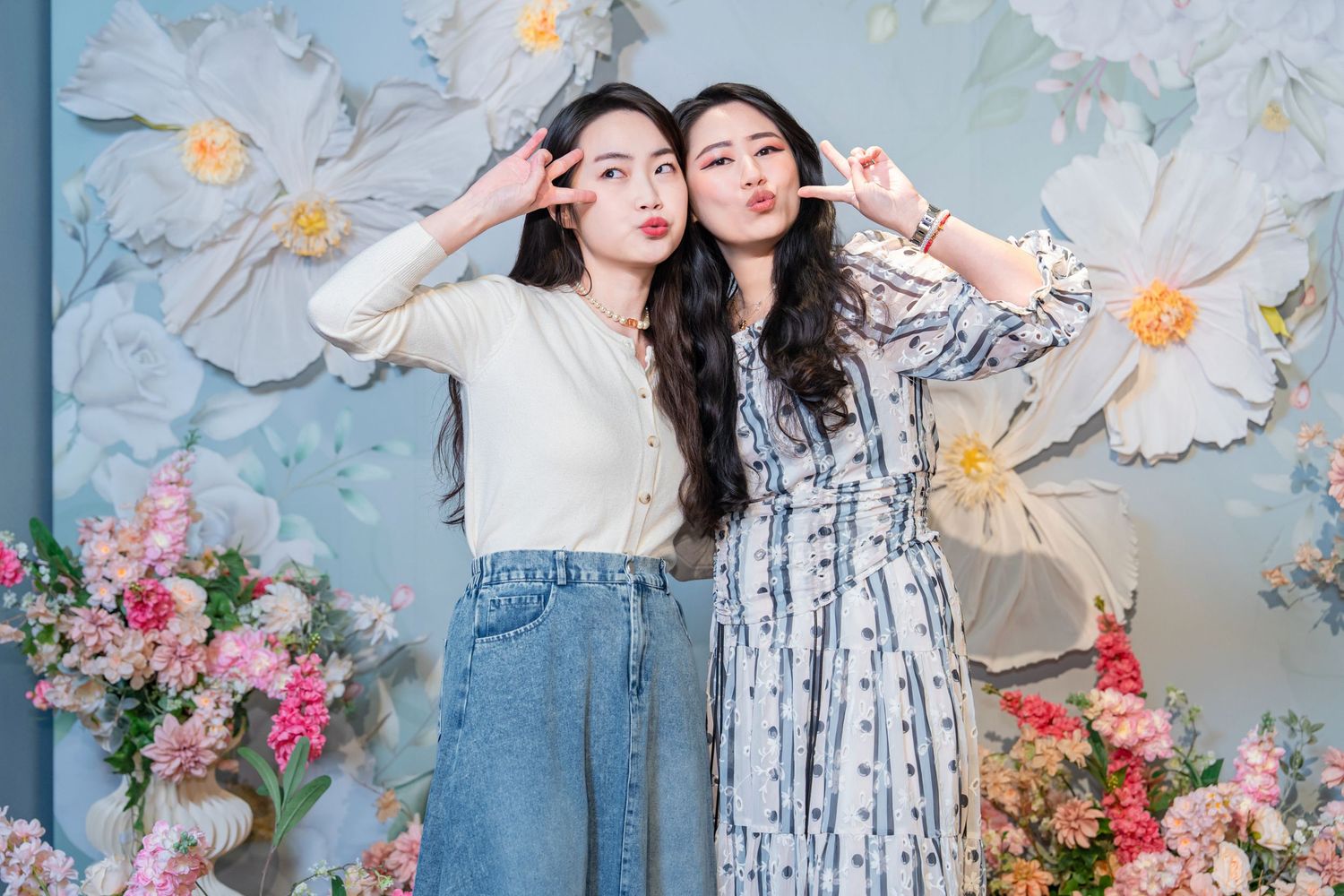 Two people in matching blue and white striped dresses pose together against a floral backdrop with white flowers.