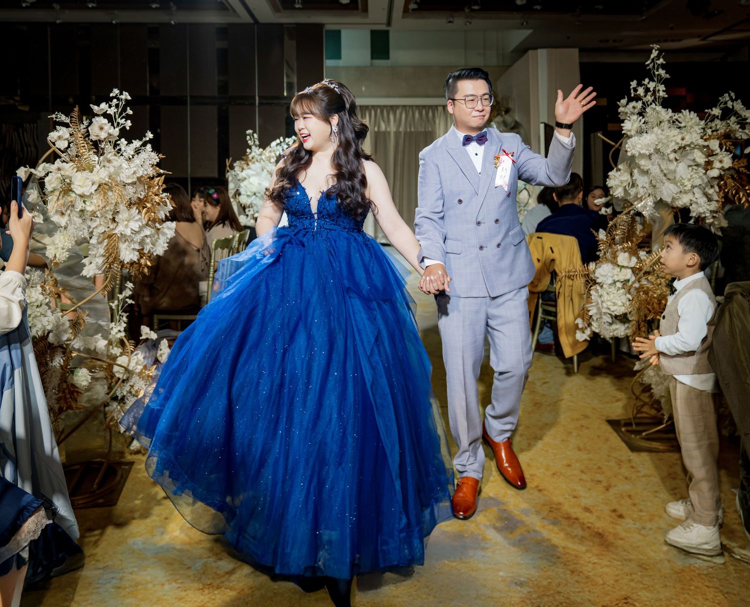 A couple in a blue ballgown and light gray suit dance together at their wedding reception surrounded by floral decor.